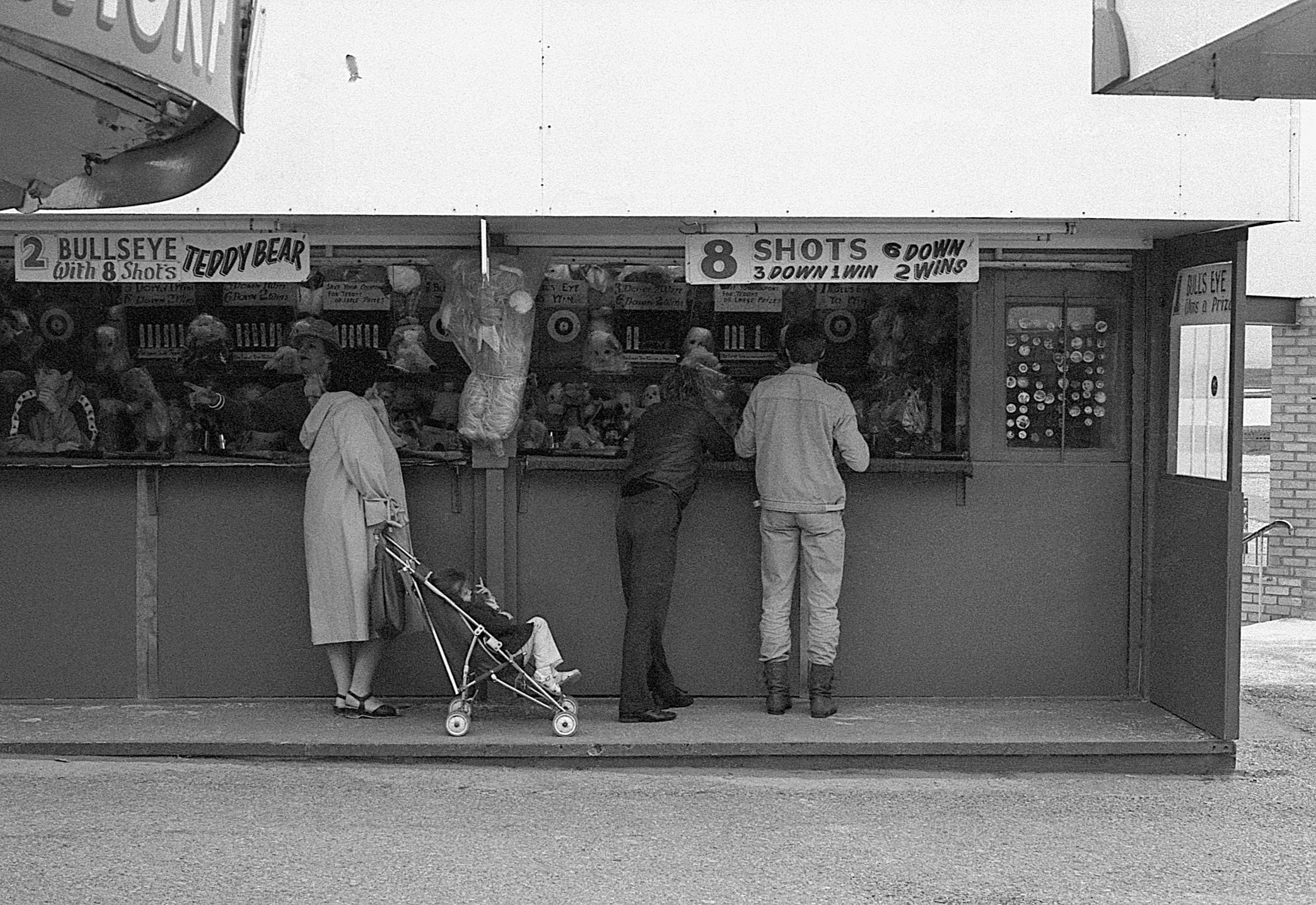A black-and-white photo of a carnival game booth with signs for shooting targets. Several people are playing or waiting in line, including a woman with a stroller, a man and a woman chatting at the counter.