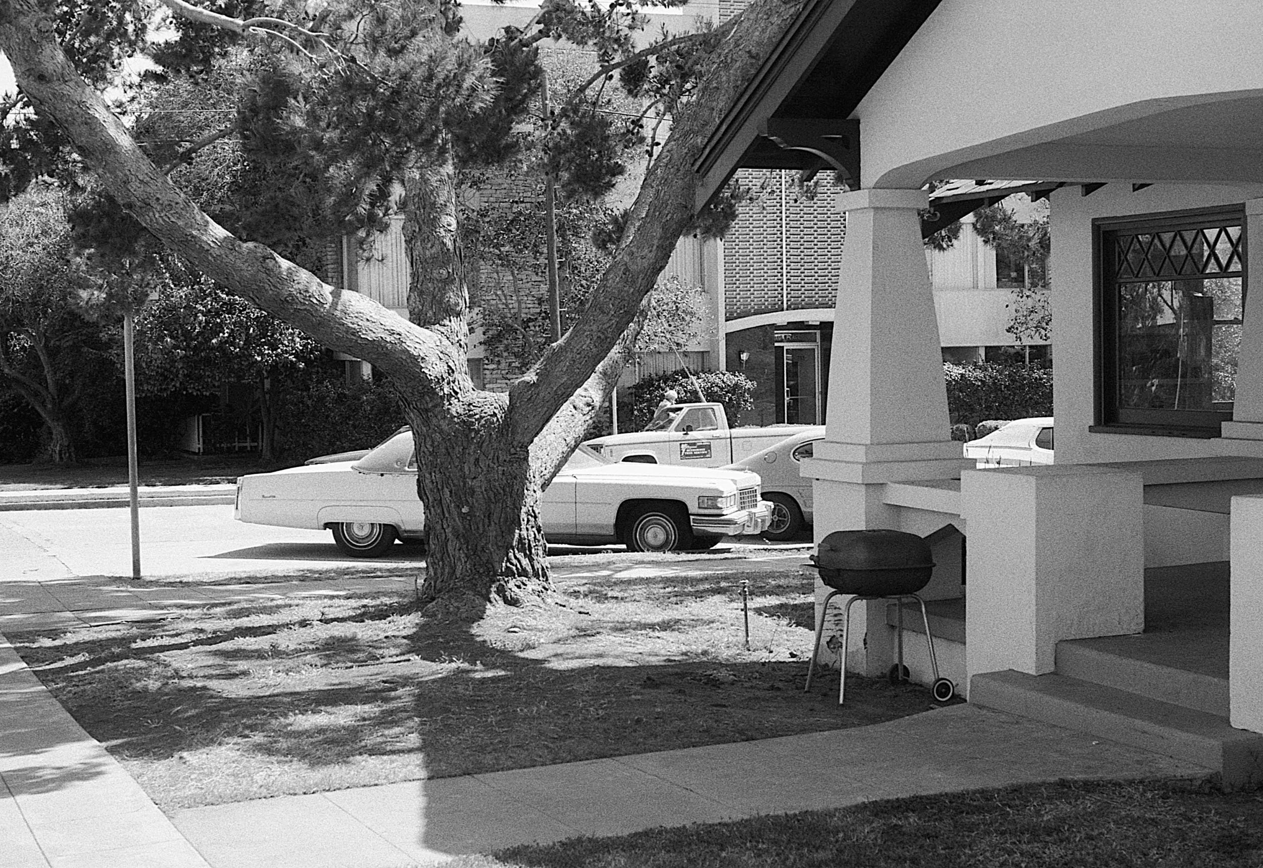 Black and white photo of a residential street view with a large tree in the foreground, a classic car parked on the street, a house porch with a grill, and another car in the background.