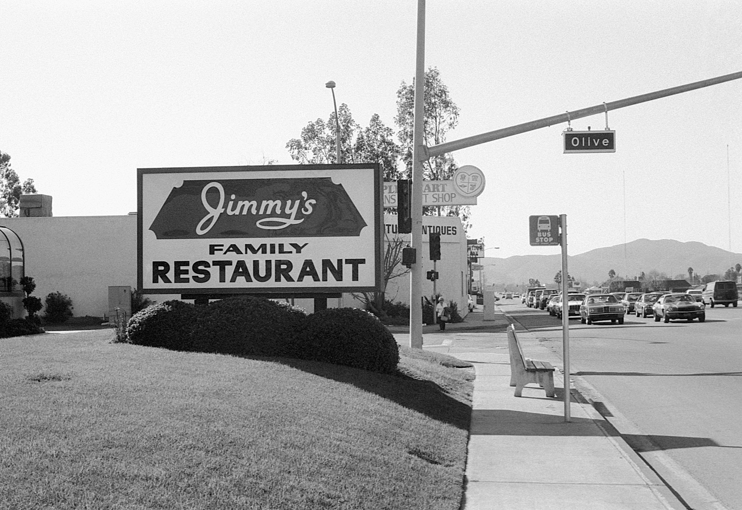 A sidewalk beside a parking lot with cars, a bus stop sign, and a street sign with the name 'Olive'. There are trees, mountains in the background, and a large sign for 'Jimmy's Family Restaurant' in the foreground.