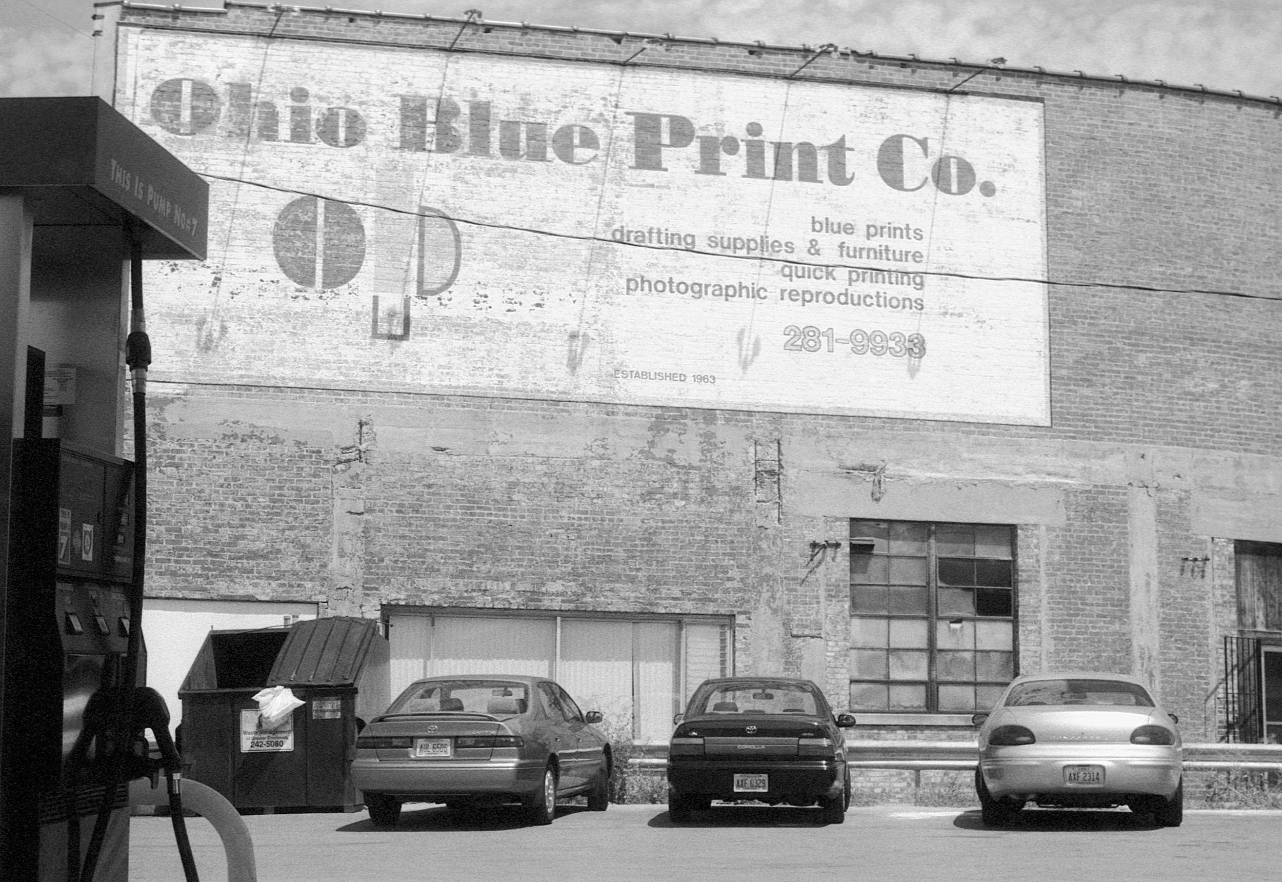 A black and white photo of a brick building with a large sign that reads 'Olio Blue Print Co.' advertising photo printing, drafting supplies, and furniture, with a phone number 281-9933. There are three parked cars and a person at a vending machine o