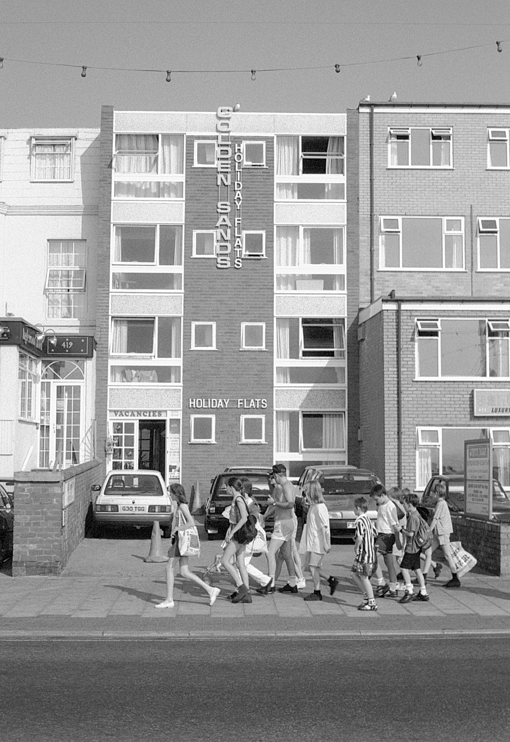 A group of children and a woman crossing the street in front of a hotel building with a sign that reads "Holiday Flats" and "Vacancies." The building has multiple windows and a parking lot with cars in front.