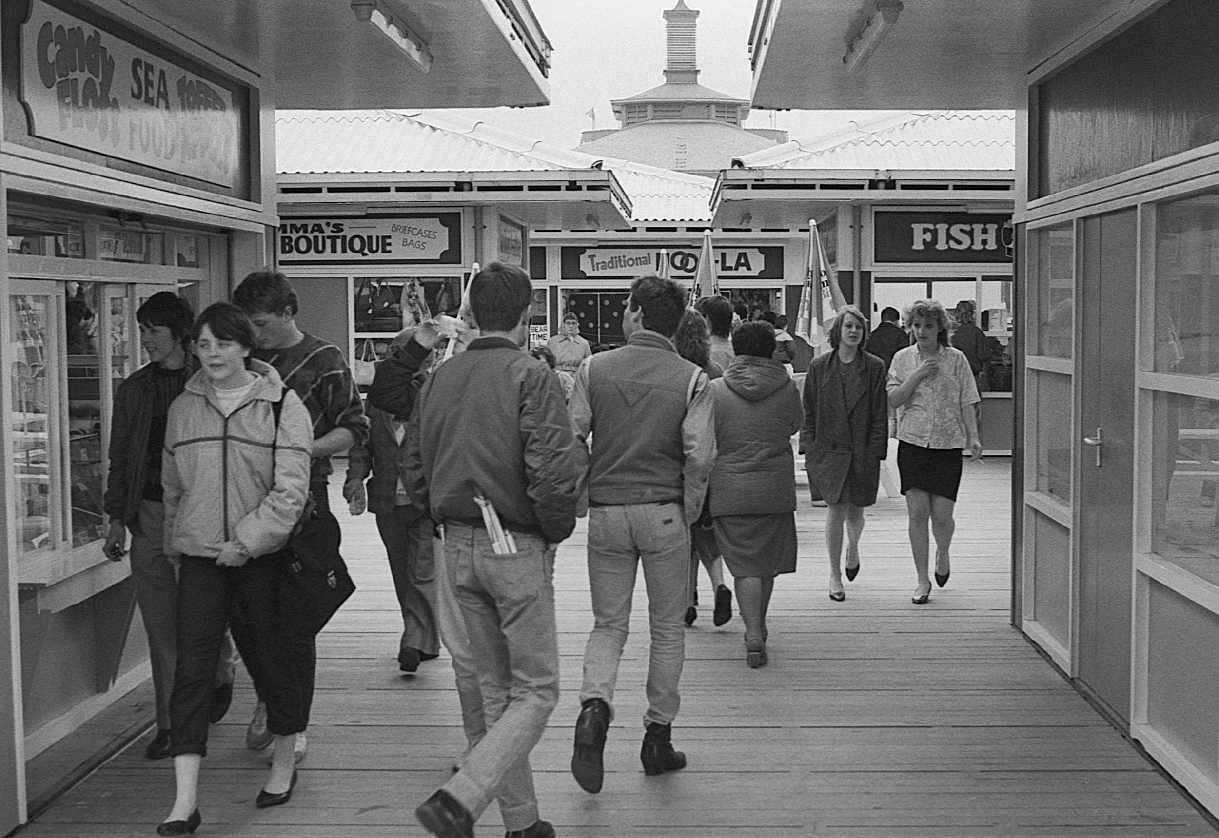 Black and white photo of people walking in a marketplace or promenade, with storefronts for seafood and fish in the background.