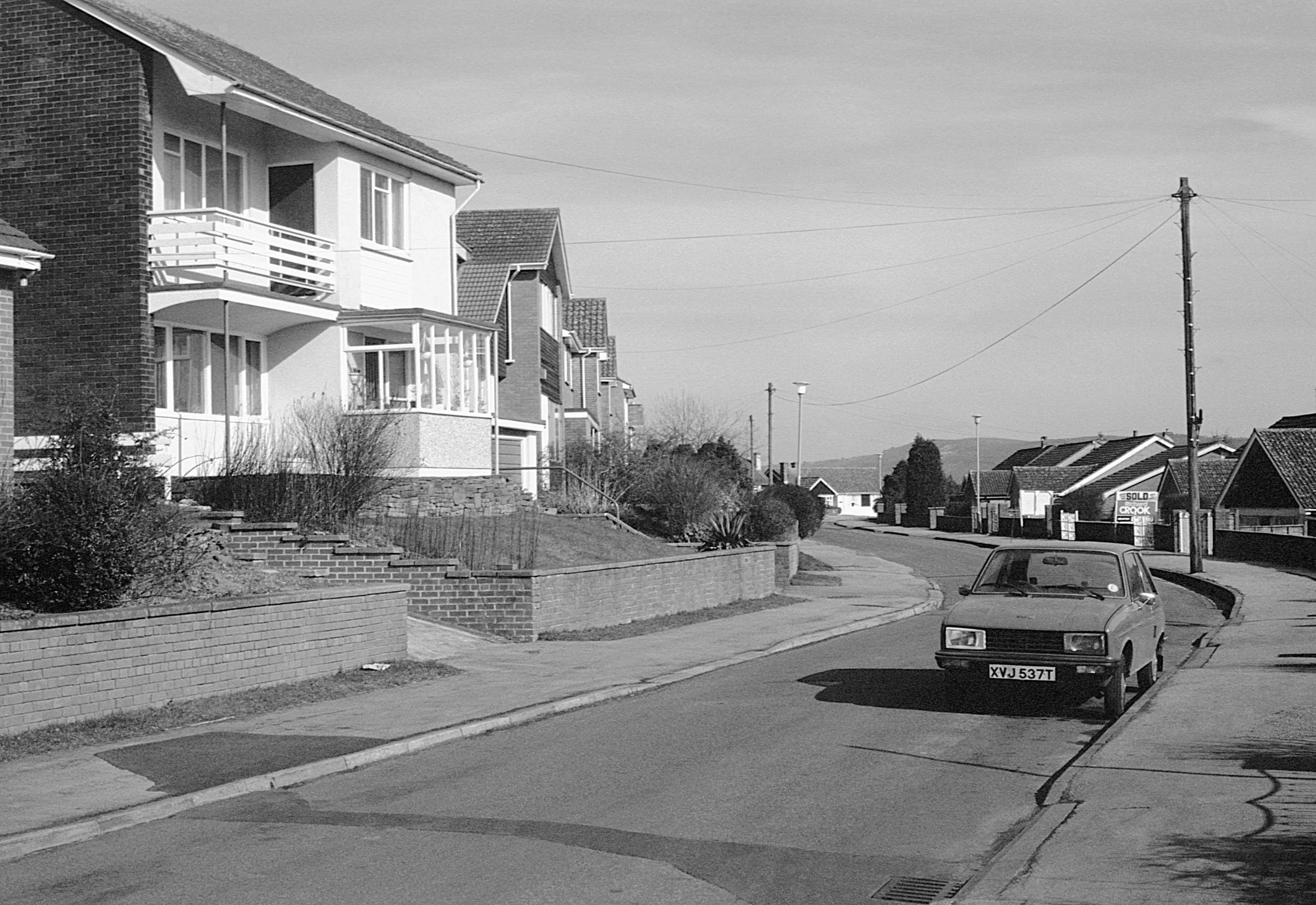 A black and white photograph of a suburban street with houses, shrubs, and a parked car on the curb.