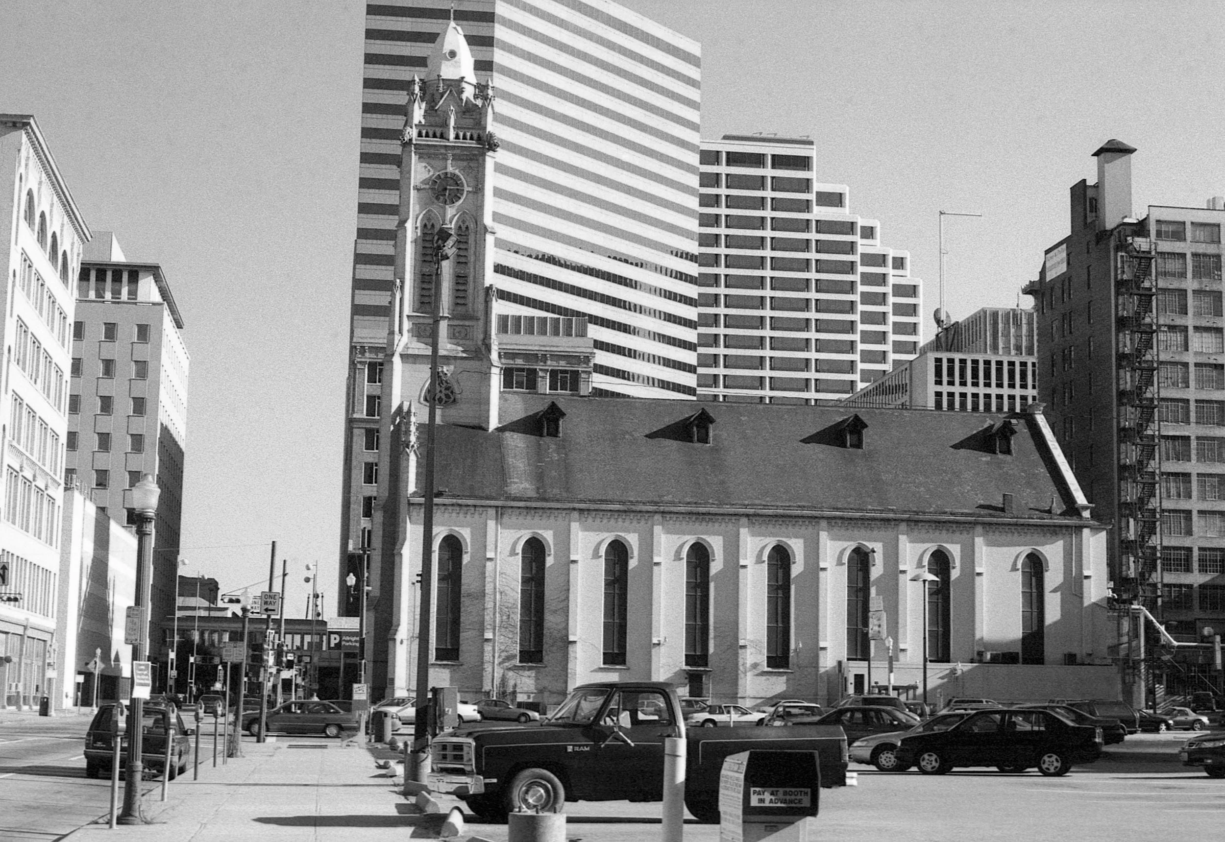 Black and white photo of a city scene featuring a historic church with a tall clock tower in the foreground and modern high-rise buildings in the background. There are parked cars and a truck in front of the church and a sidewalk with street lamps.