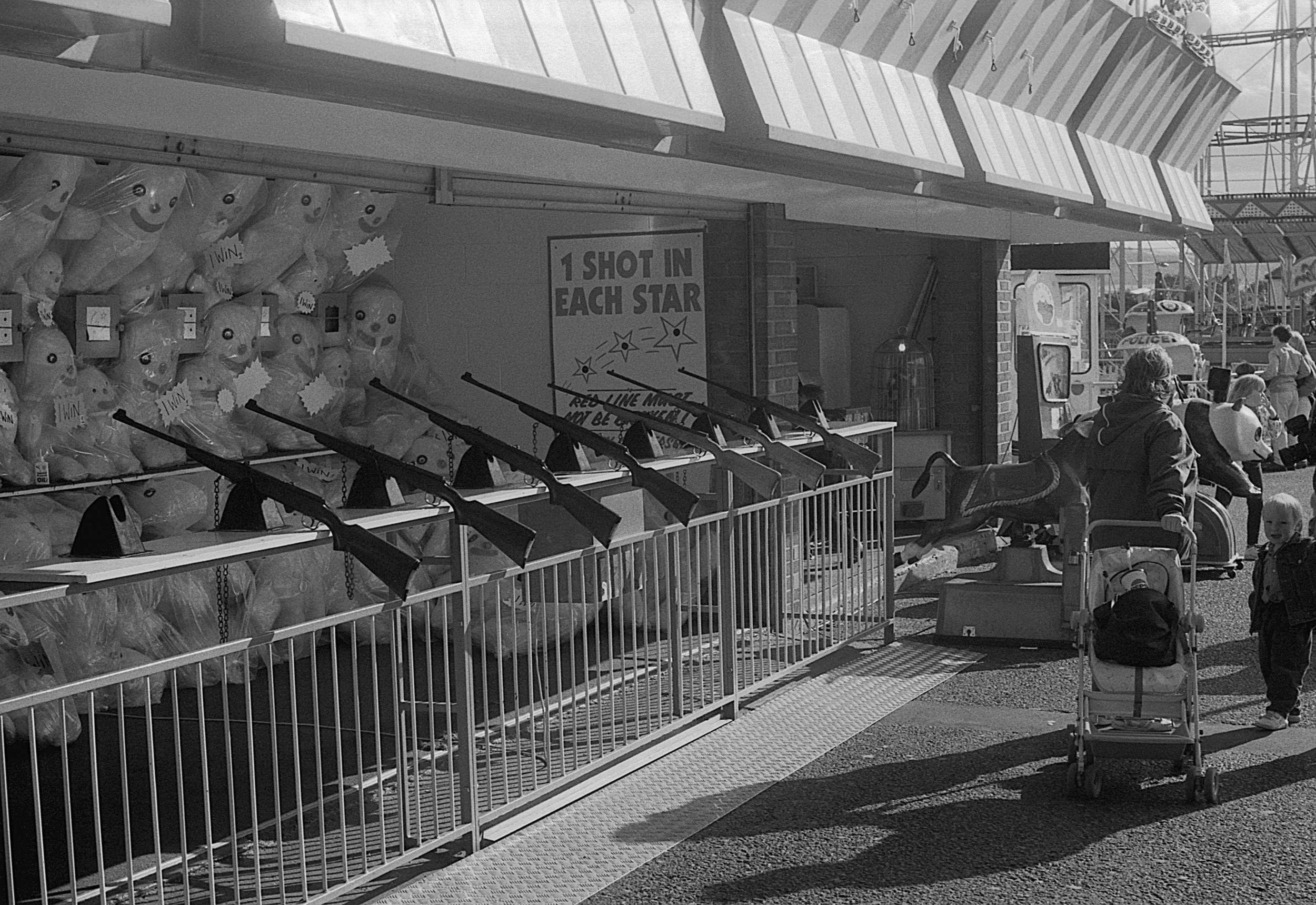 At an amusement park or arcade, a row of water guns are set up for a shooting game, with plush toys and targets in the background. People, including children and adults, are visible enjoying the attractions.