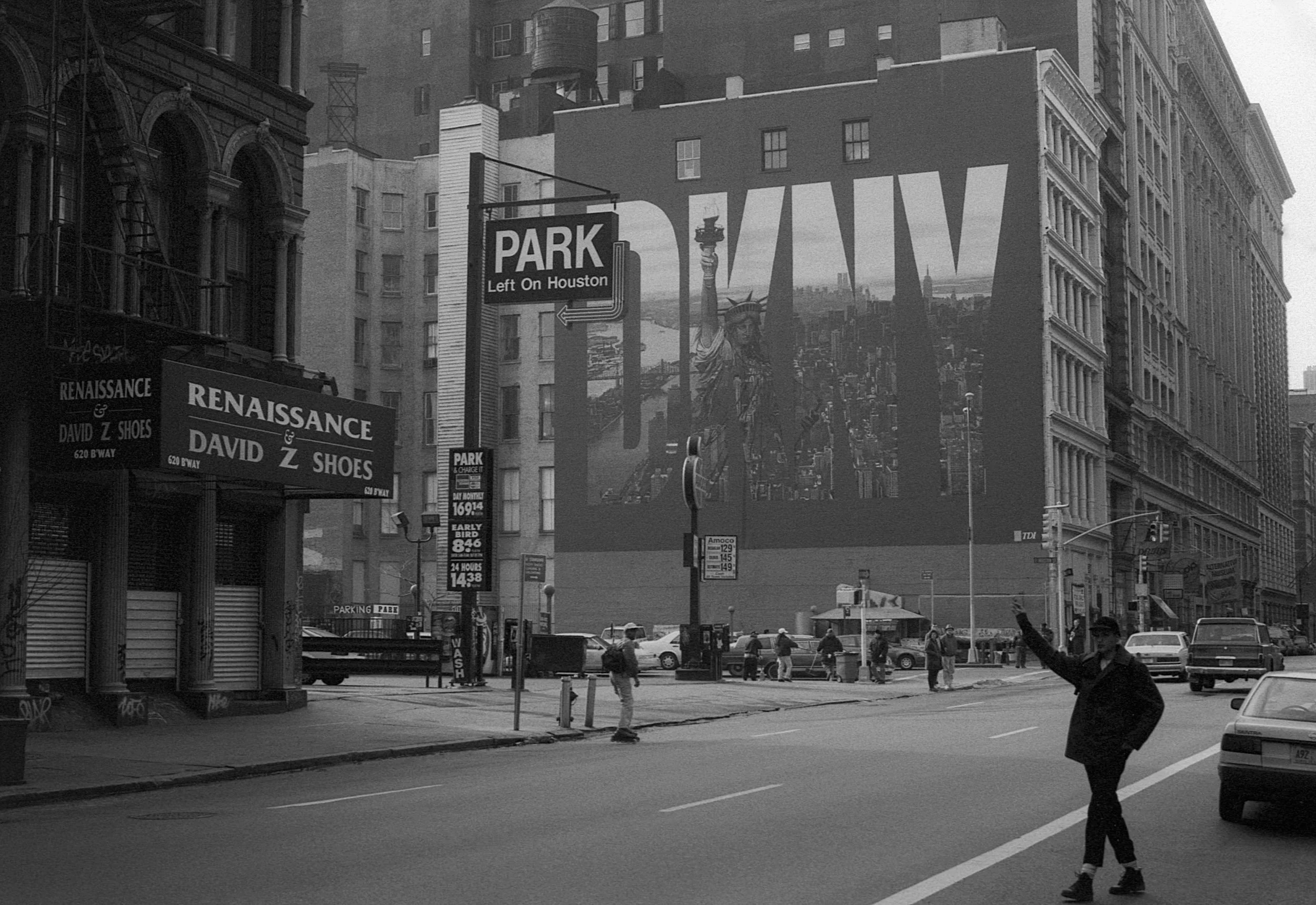 A street scene in New York City in black and white, showing pedestrians crossing the street, parked cars, and large buildings. A billboard features the Statue of Liberty with a cityscape in the background. There are signs for parking and a shoe store