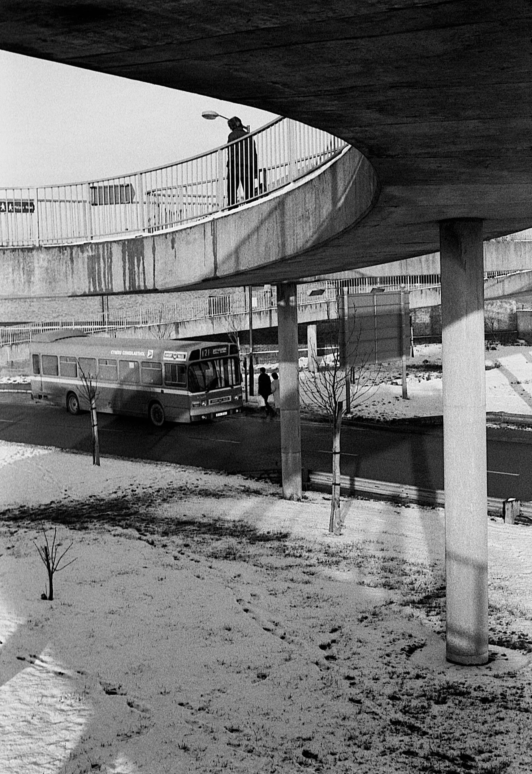 Black and white photo of an urban scene with a bus parked on the street beneath a concrete overpass. Two people are standing beside the bus, and a pedestrian is walking nearby. The ground is covered with snow, and there are a few small, leafless tree