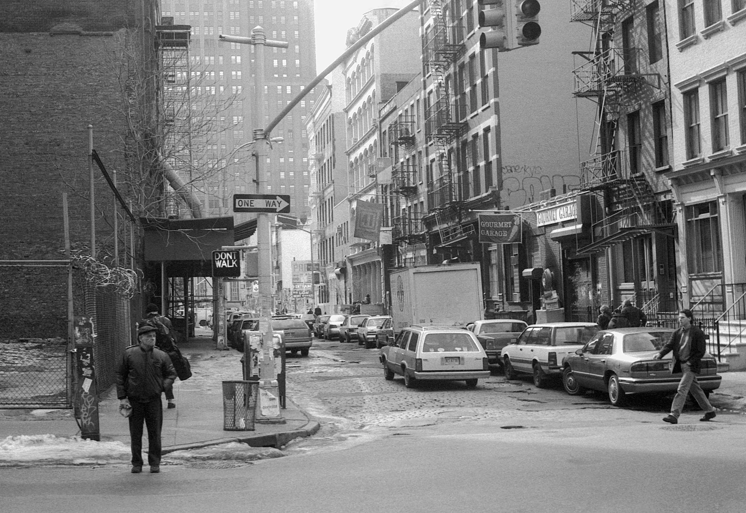Black and white street scene with parked cars, pedestrians, and buildings. Traffic signs indicate one-way and no walking zones. Some buildings have fire escapes, and there is some graffiti and signage.