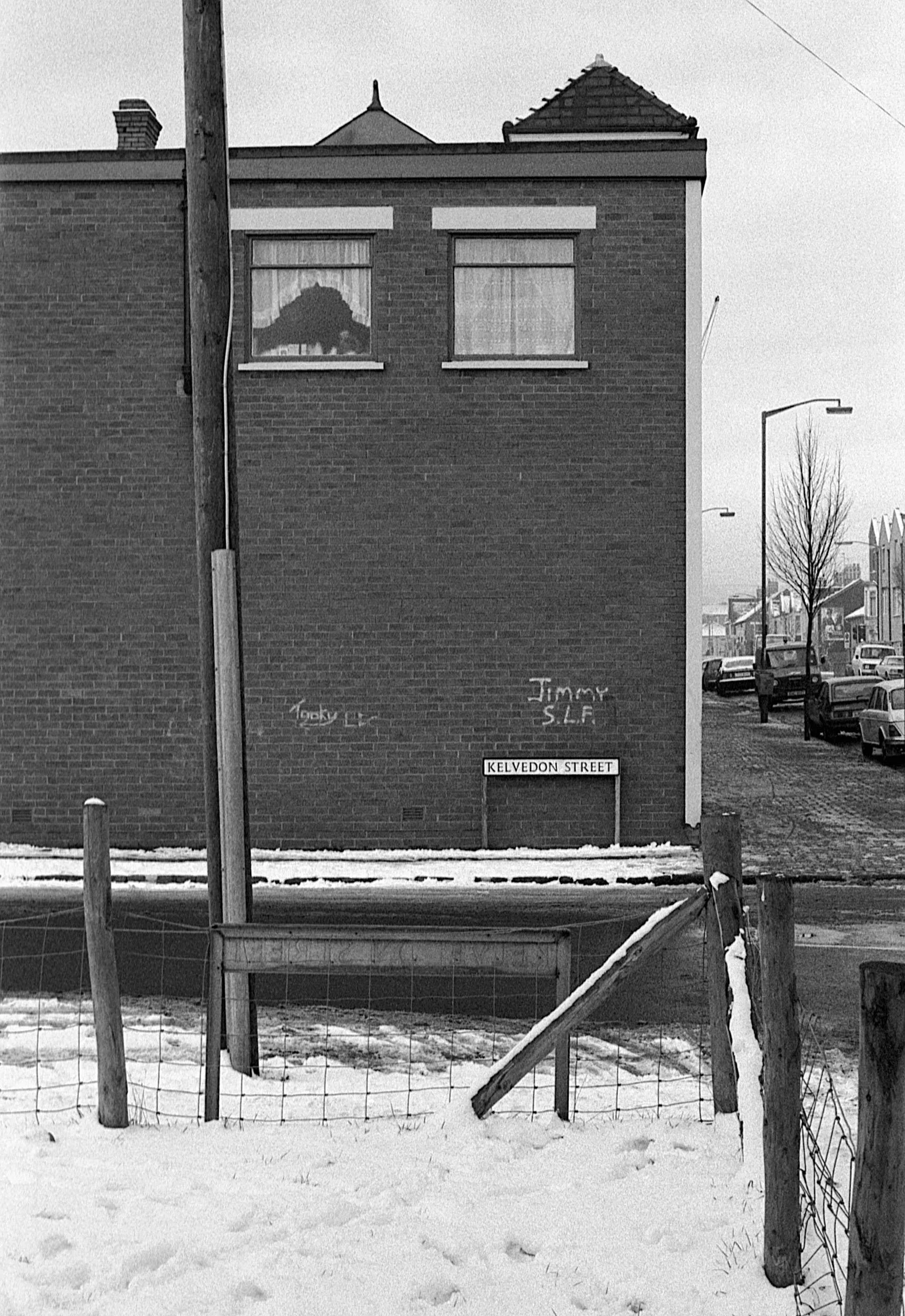 Black and white photo of a brick building with two windows and a chimney, a street sign that reads 'Kelvedon Street,' graffiti on the wall, and a snow-covered sidewalk with a fence and parked cars along the street.