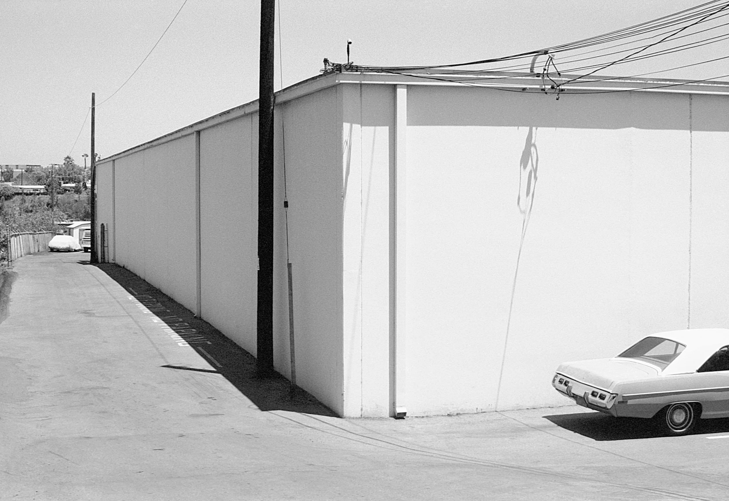 Black and white photo of a large, plain white building with a flat roof, a car parked to the right, utility poles, and power lines overhead.