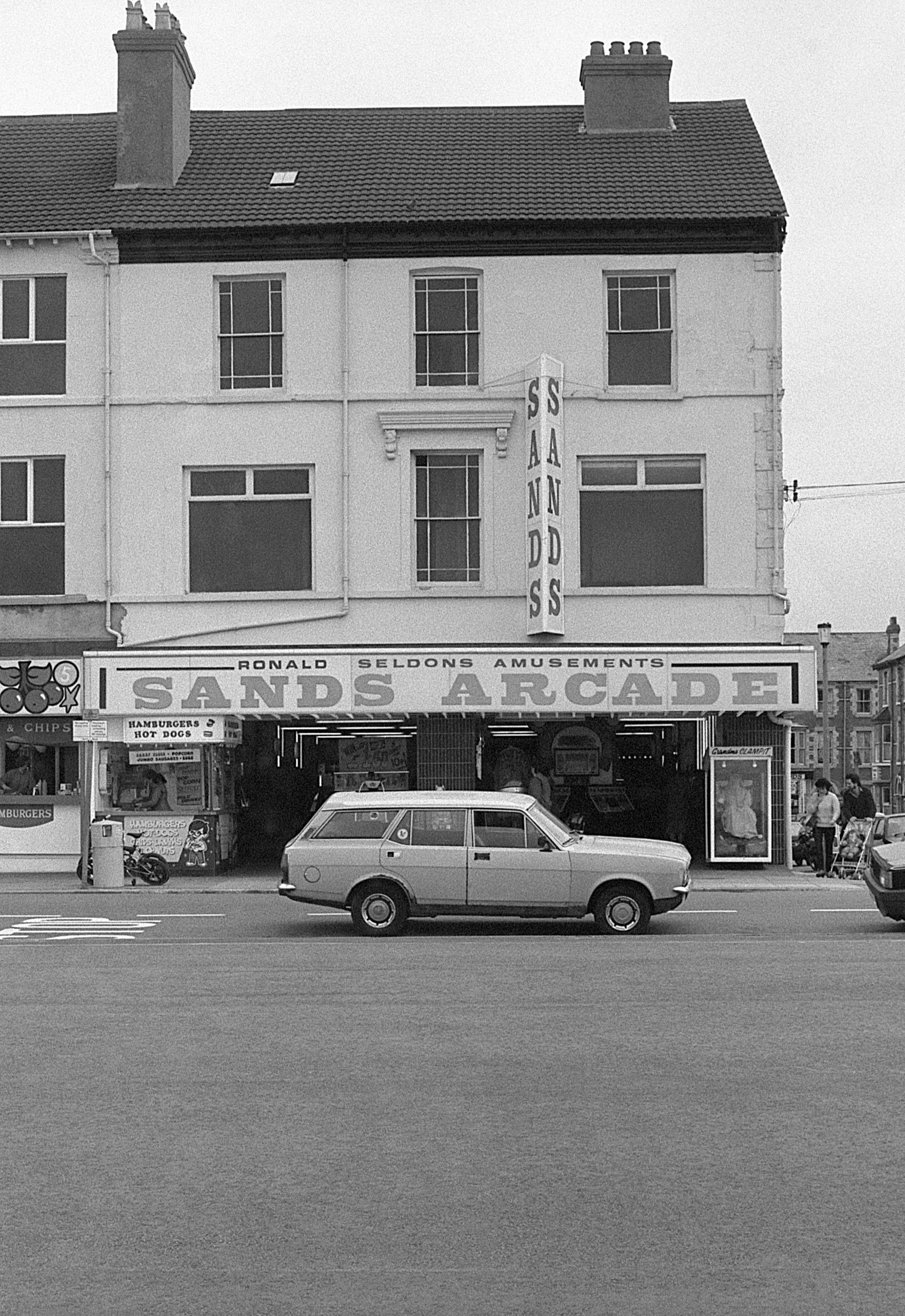 Black and white photo of Sands Arcade, a historic amusement arcade with a sign displaying 'SANDS' vertically and 'SANDS ARCADE' horizontally. The building has three stories with windows, and a car is parked on the street in front of it. There are peo