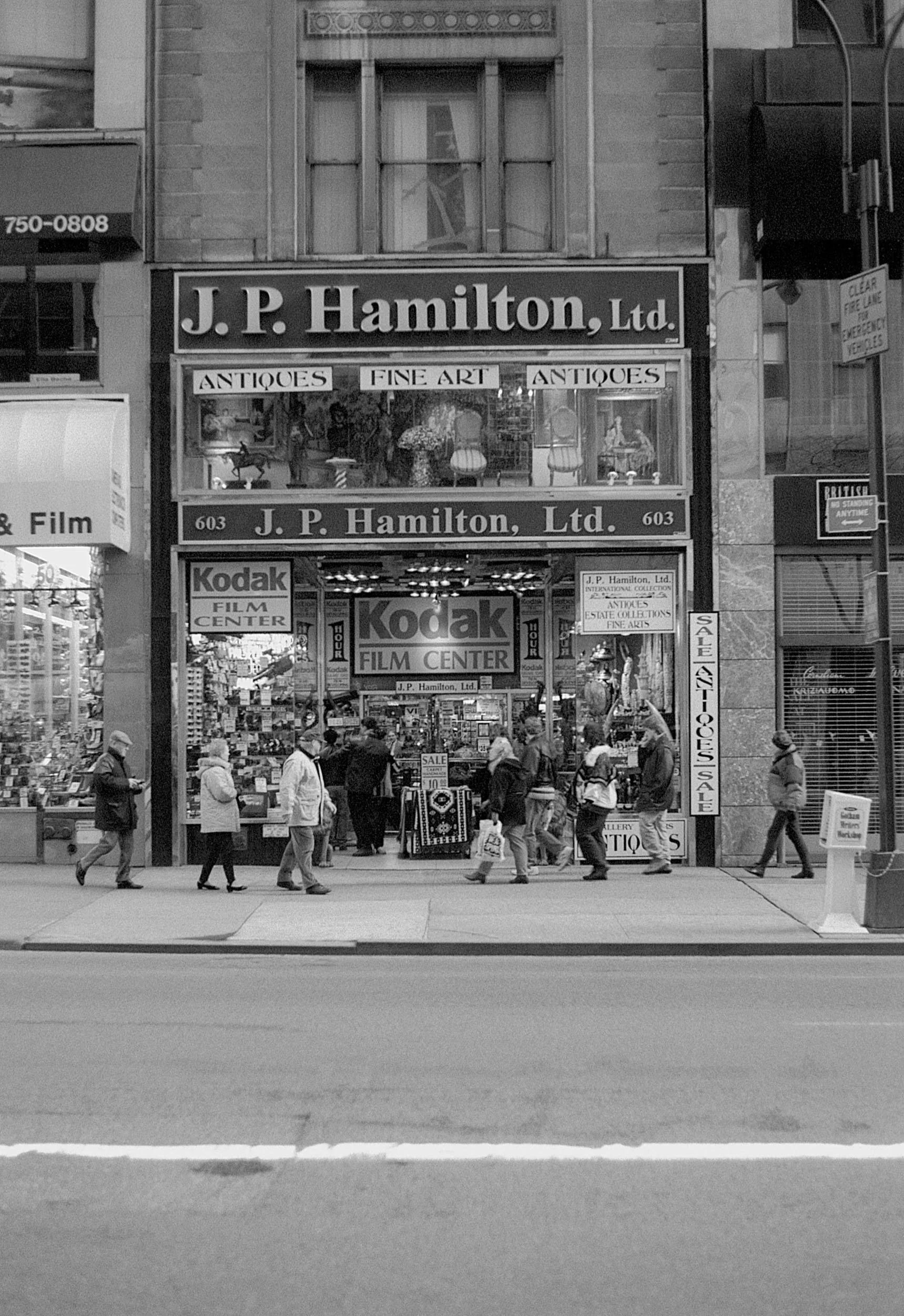 A storefront for J. P. Hamilton Ltd. featuring antique and art items, with a Kodak film center inside. Pedestrians walk past the store on a city sidewalk, some carrying shopping bags, in a black and white photo.
