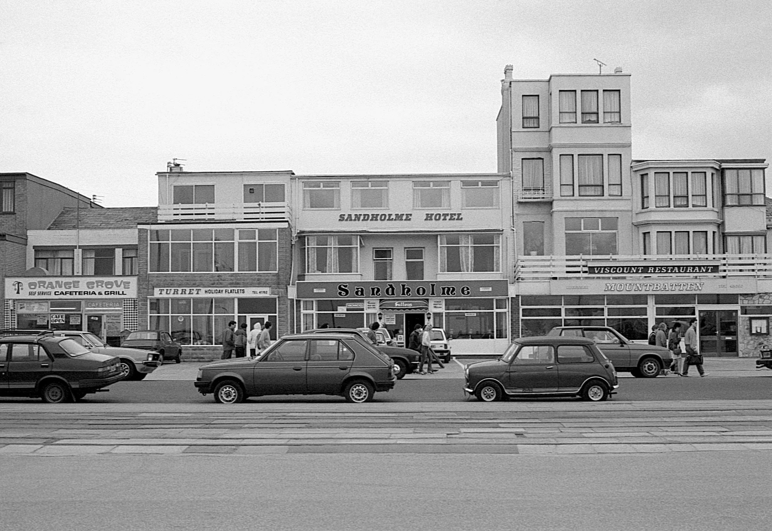 Black and white photo of a street with parked cars in front of buildings, including a hotel and restaurants, with people walking along the sidewalk.