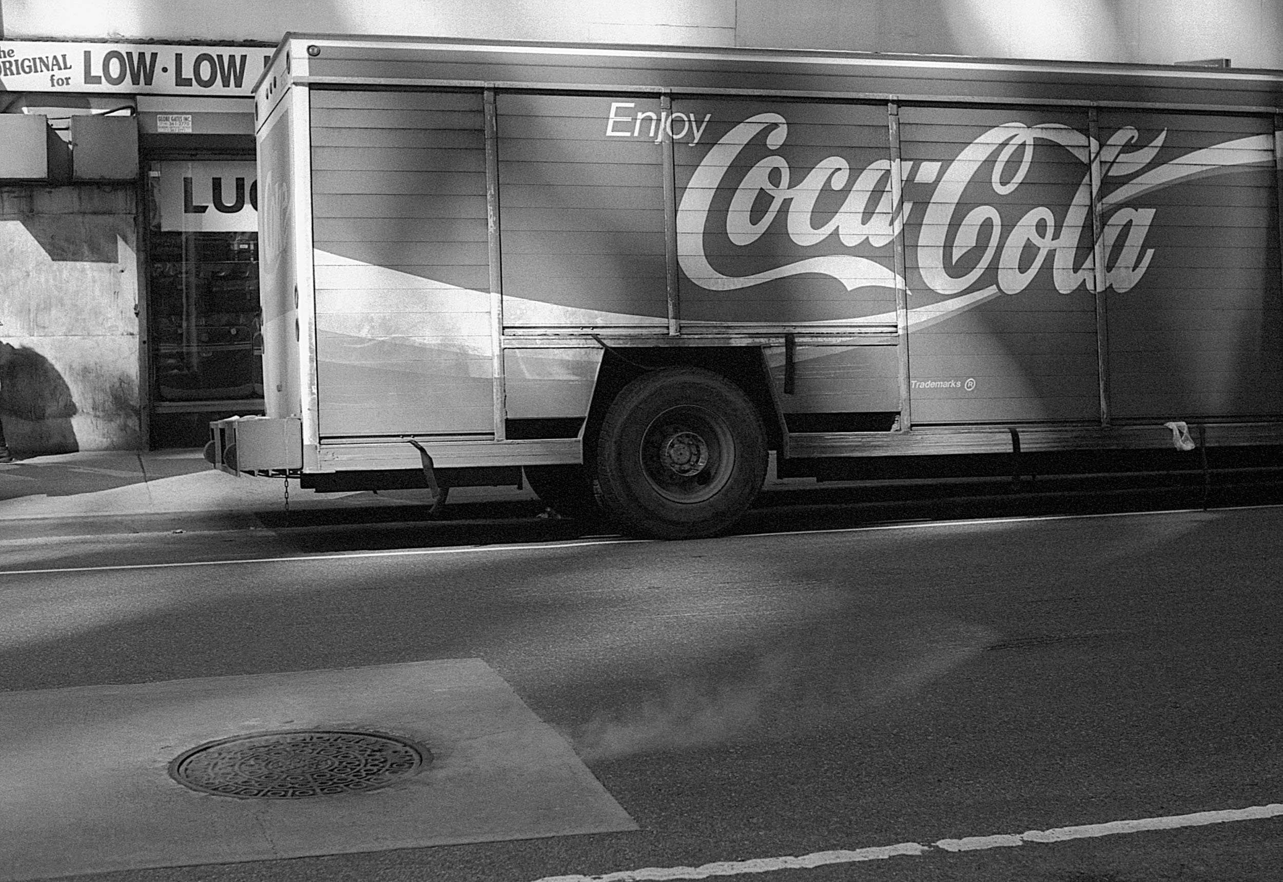 A black and white photo of a Coca-Cola delivery truck parked on a street. The truck has the Coca-Cola logo on its side and is adjacent to a building with signs that read 'Low Low' and other partial text.