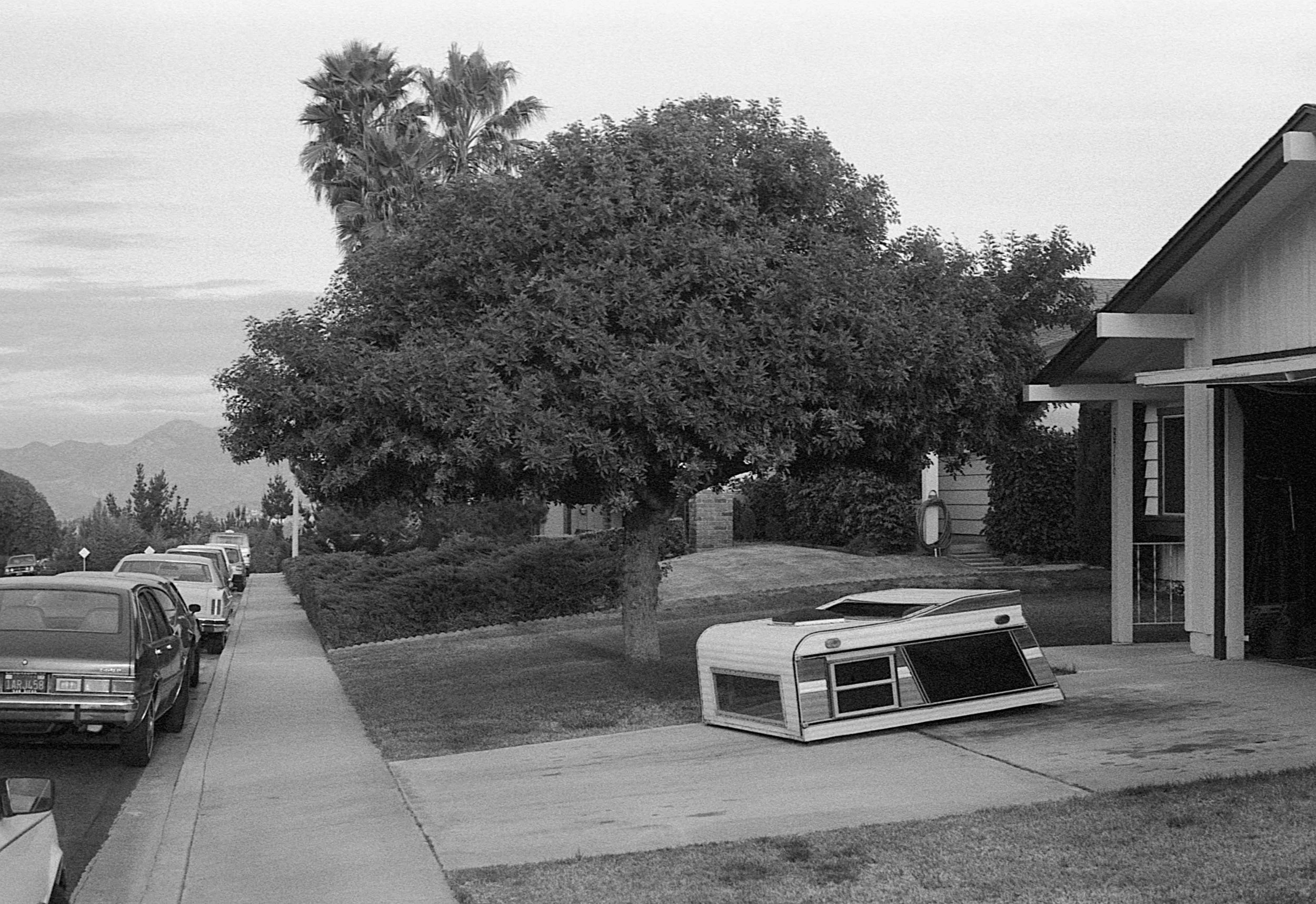 A residential street with parked cars along the curb, a large tree in the front yard, and a fallen RV roof section on the driveway in front of a house.