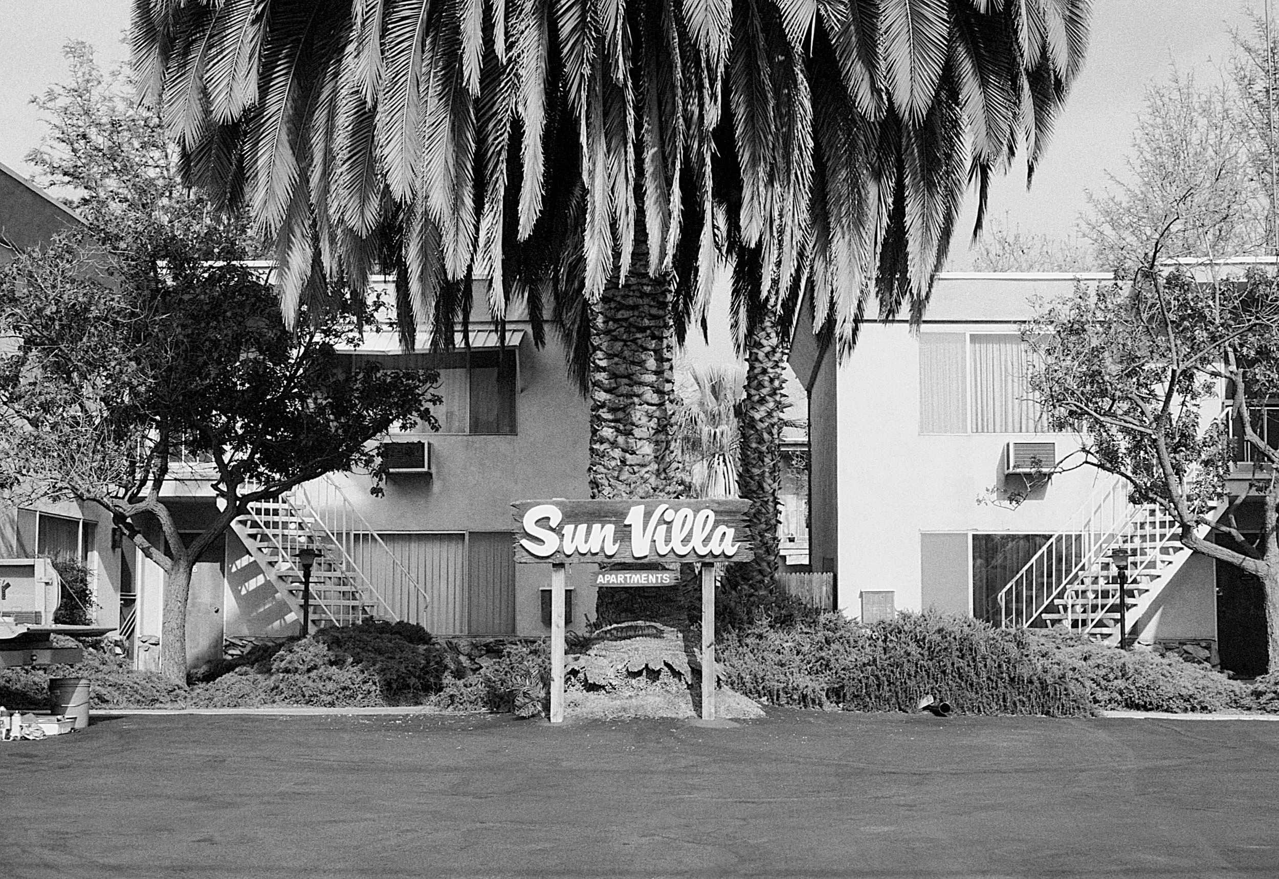 Black and white photo of an apartment complex with a sign that reads 'Sun Villa Apartments' in front, surrounded by tall palm trees and smaller trees, with stairs leading up to the apartments.
