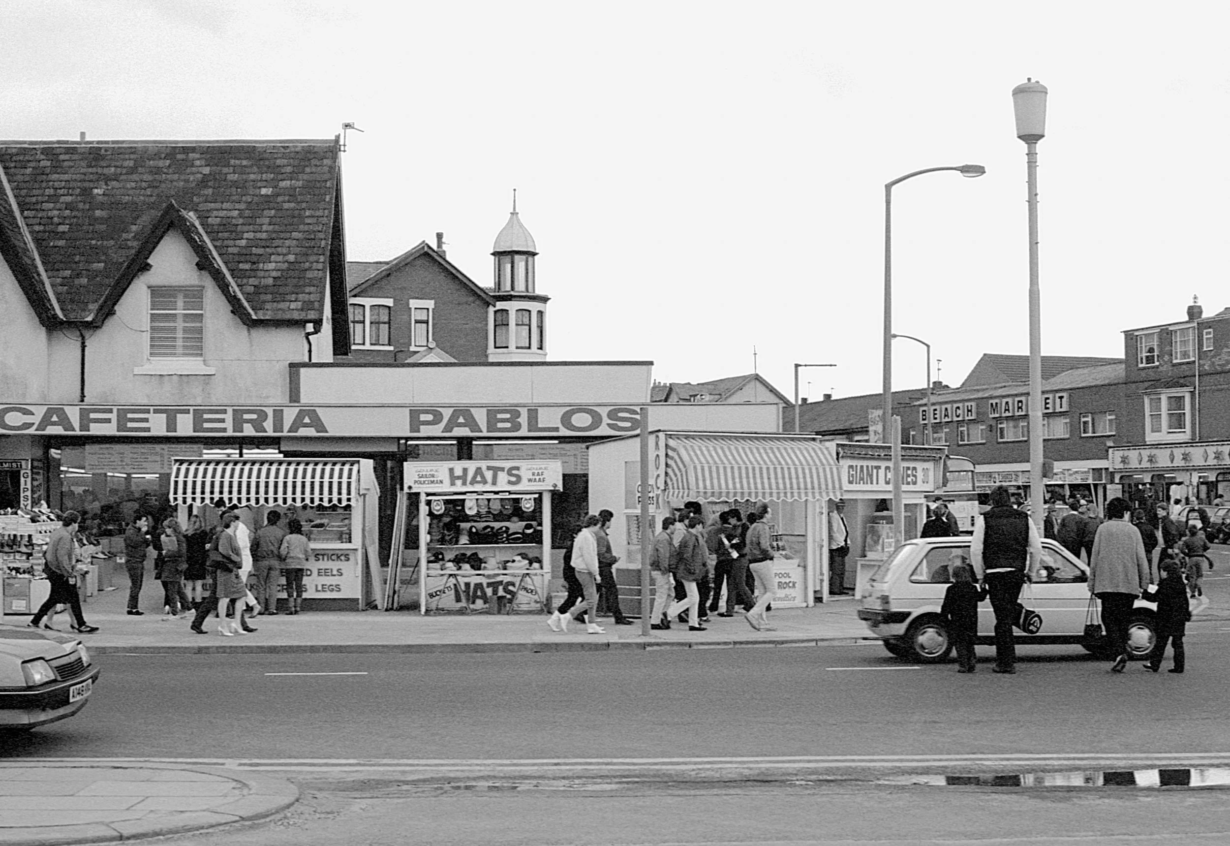 Black and white photo of a busy street scene with people walking on the sidewalk, small stalls selling hats, and storefront signs that read 'CAFETERIA PABLOS,' 'GIANT GAMES,' and 'BEACH MARKET.' There are street lamps and buildings in the background.