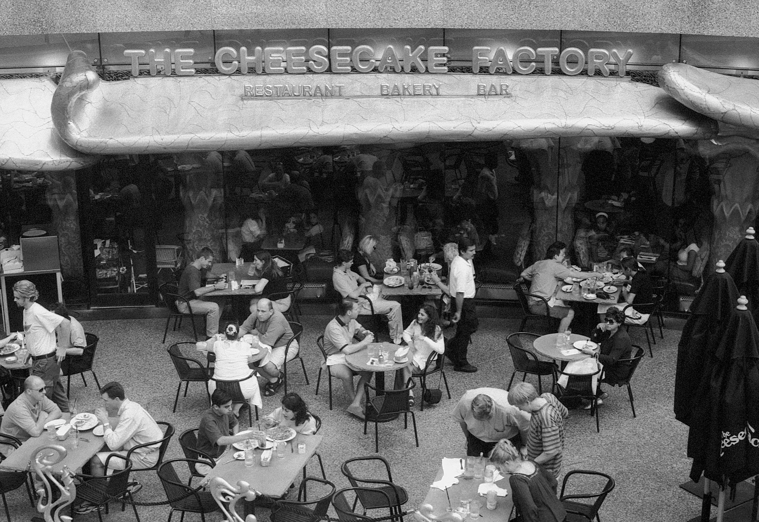 People dining inside The Cheesecake Factory restaurant, with the sign above indicating it as a restaurant, bakery, and bar.