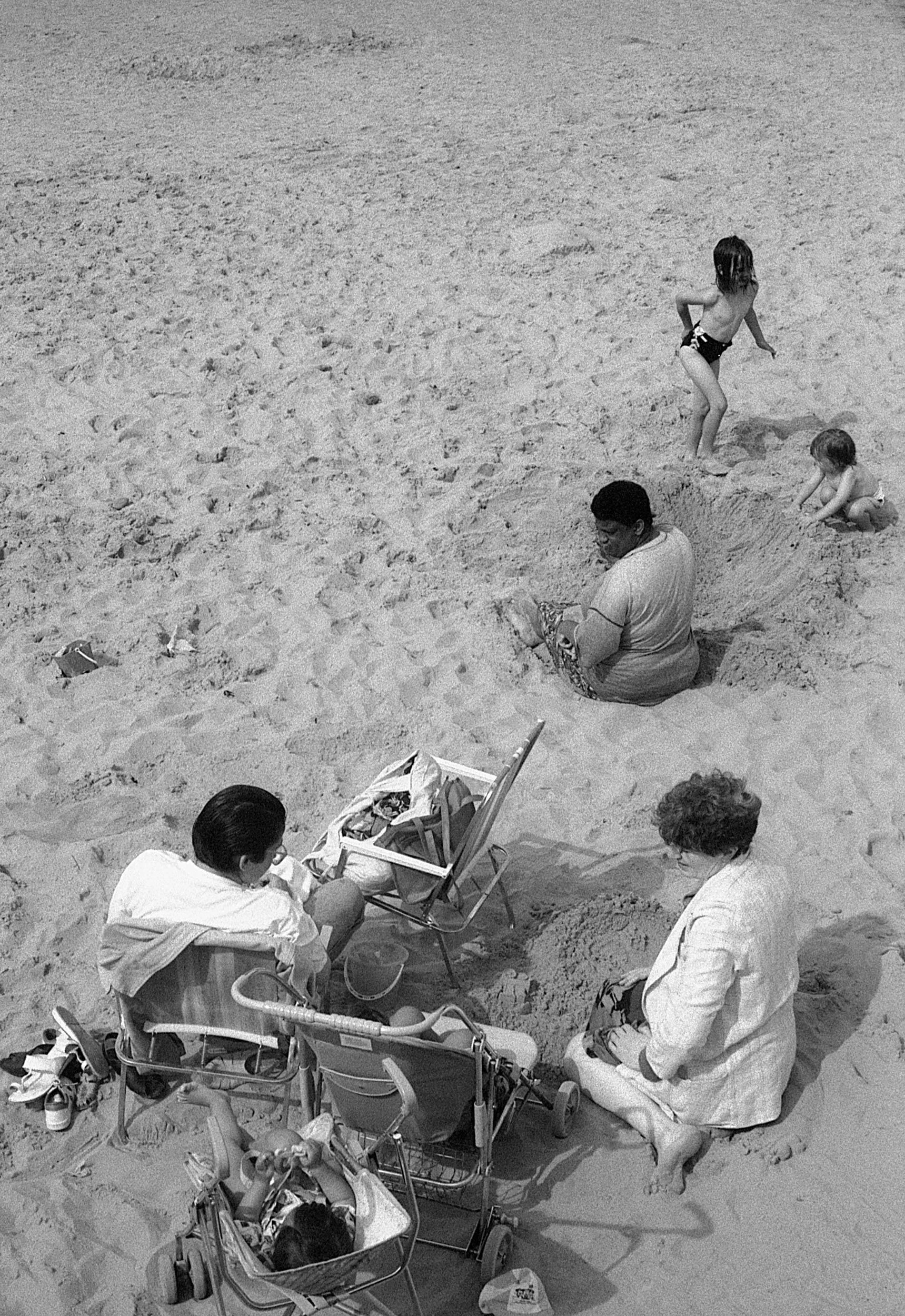 Two children and two adults on a sandy beach. One child is standing, another is crawling. Two adults are seated on the sand, one with a stroller nearby, and a bag and shoes beside them.