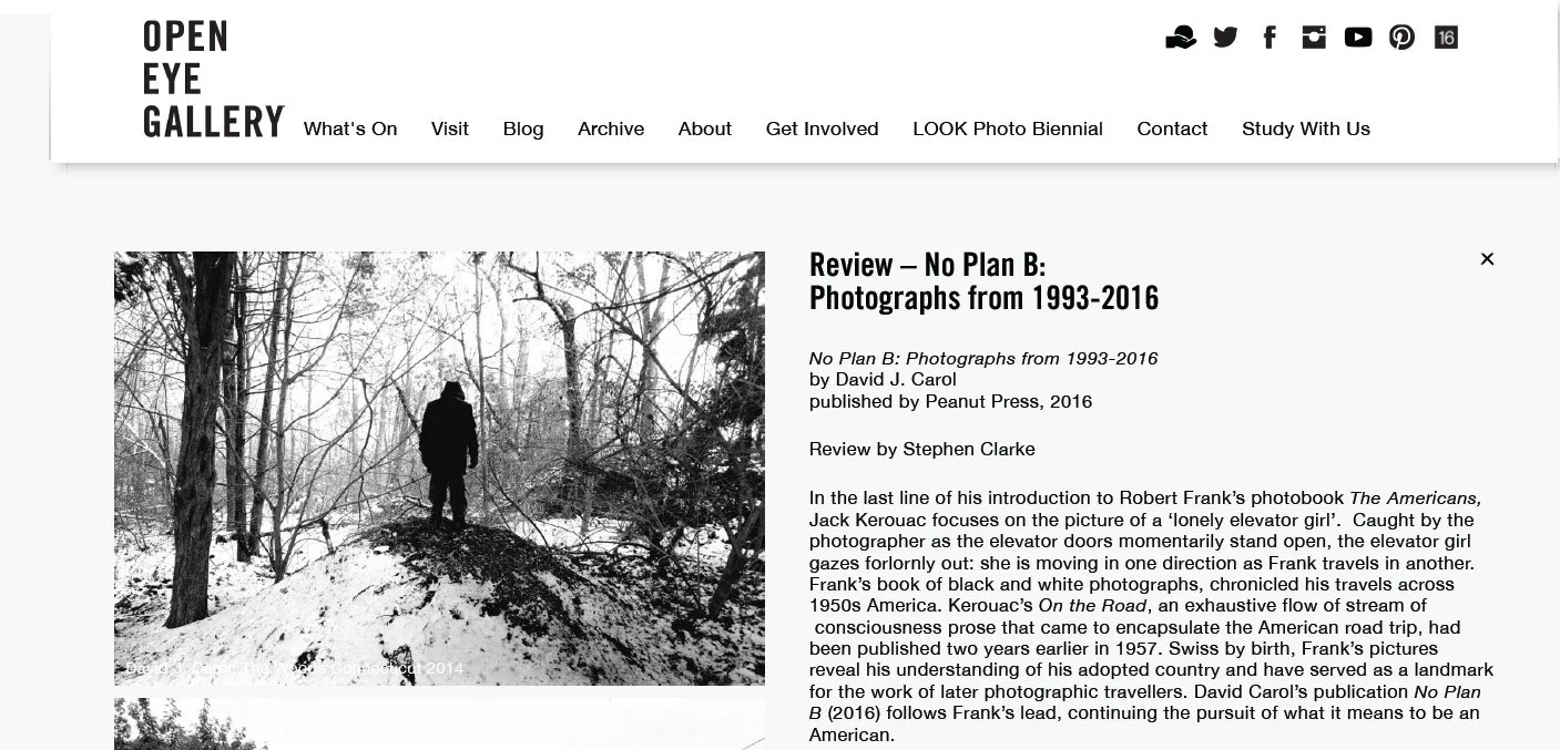 Black and white photograph of a person standing on a snow-covered hill in a forest, gazing into the distance amidst leafless trees.