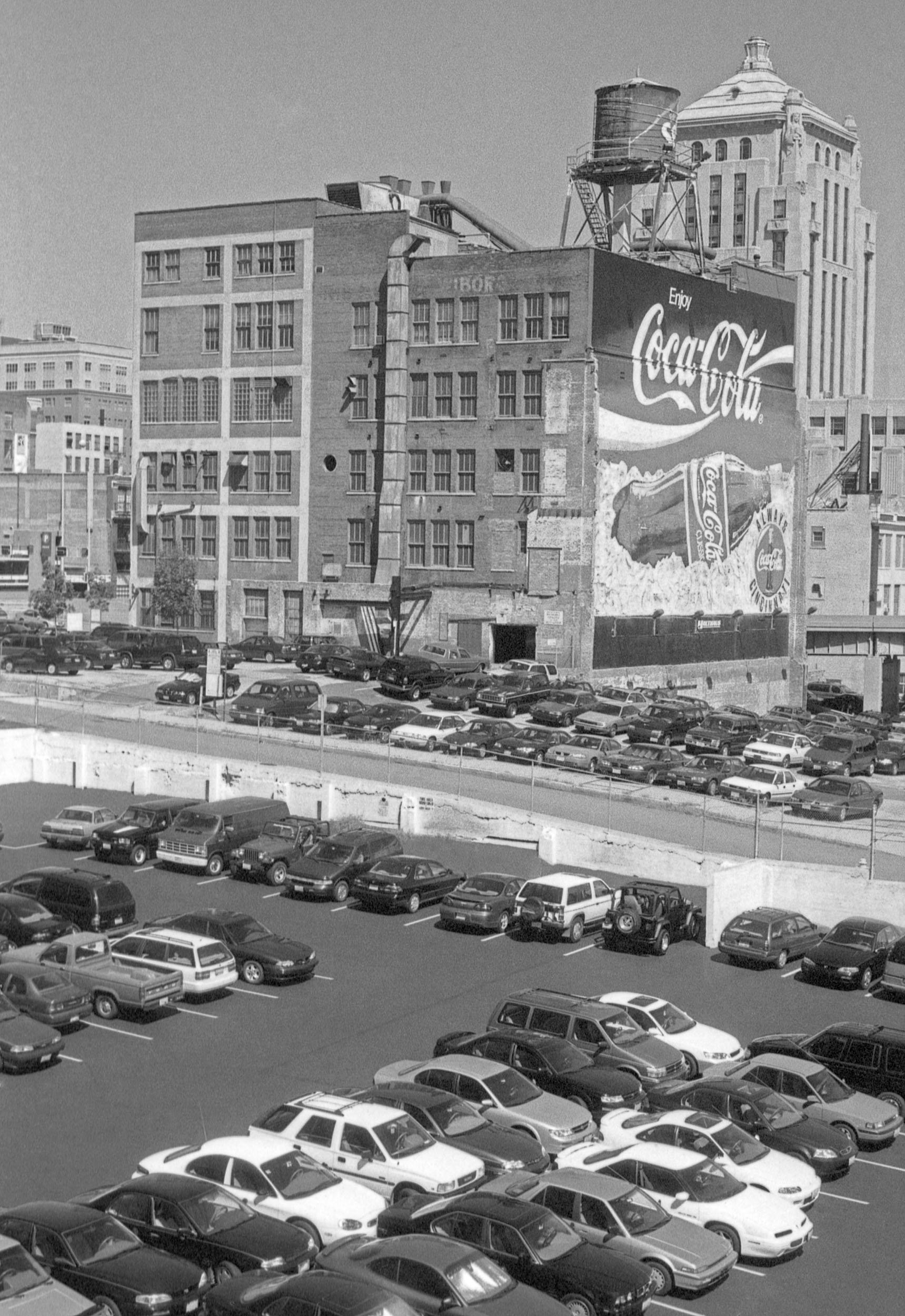 Black and white photo of a multi-story building with a large Coca-Cola advertisement on the side, parking lot with numerous cars, and cityscape in the background.