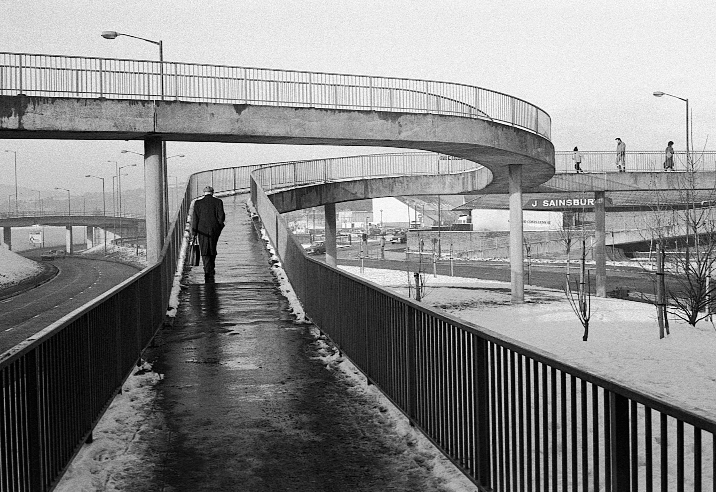 Black and white photo of a pedestrian bridge with a man walking along the sidewalk, carrying a briefcase. The bridge has winding pathways and metal railings, with a few people walking on the upper level. Snow is on the ground, and there are industria