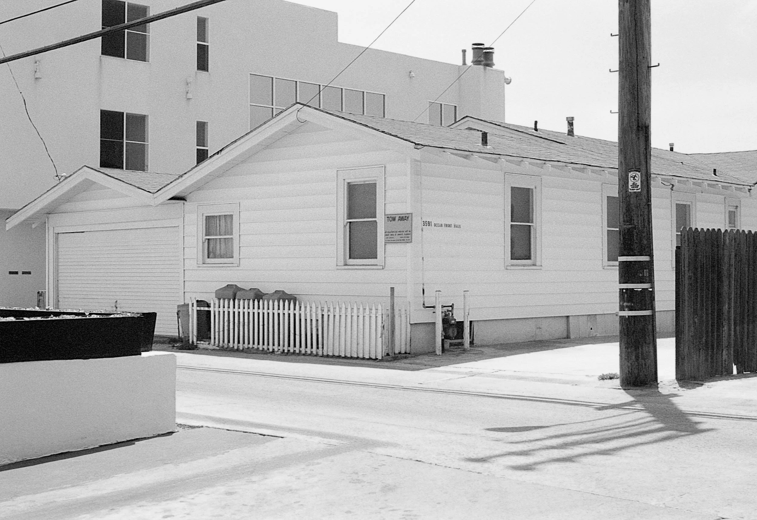 A small, white house with three windows facing the street, a white picket fence, and a garage. A large modern building is behind it. There is a street with snow and a utility pole with wires.