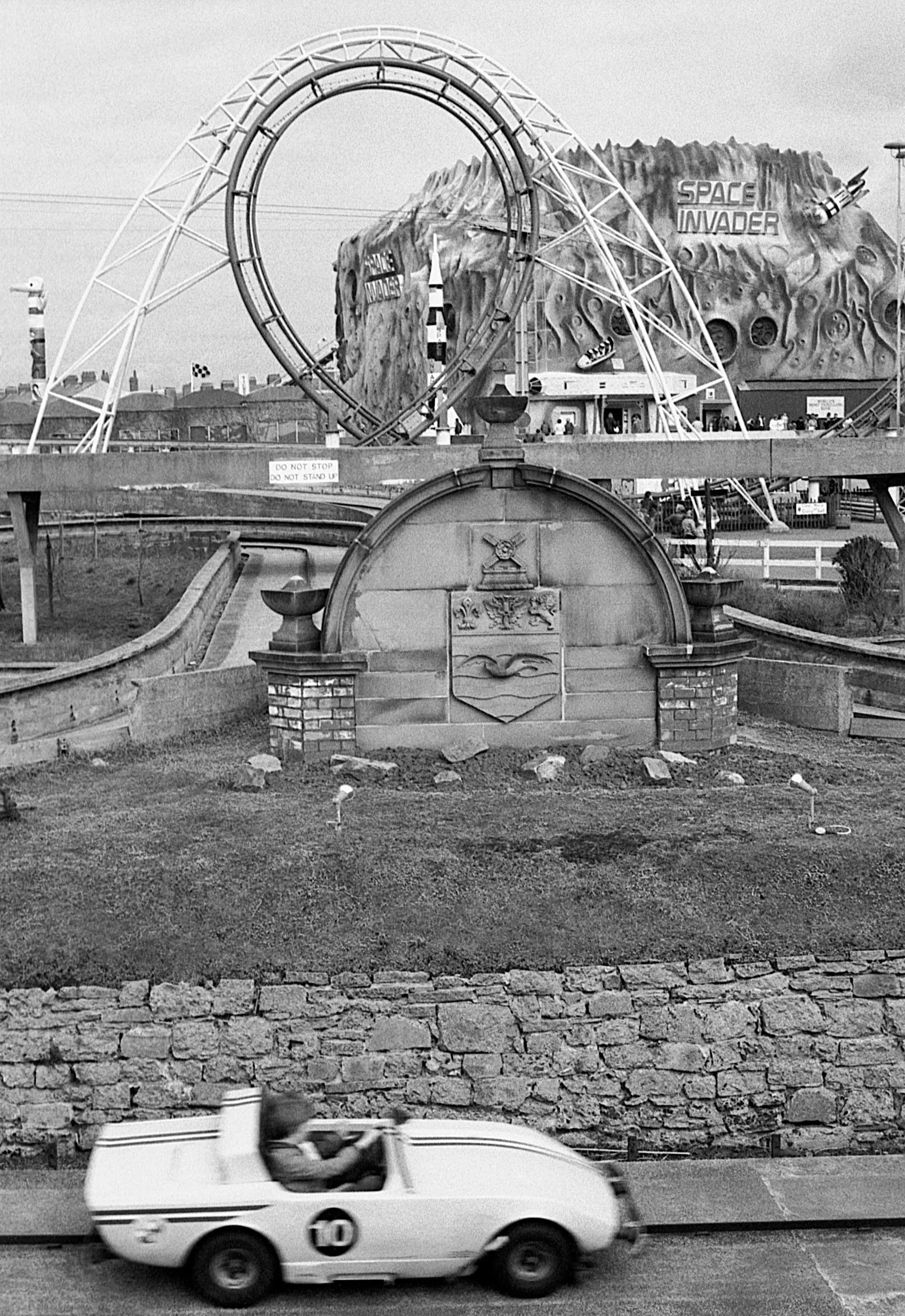 Black and white photo of an amusement park with a roller coaster named 'Space Invader' in the background. In the foreground, a vintage white race car with the number 10 on the side is driving on the street.