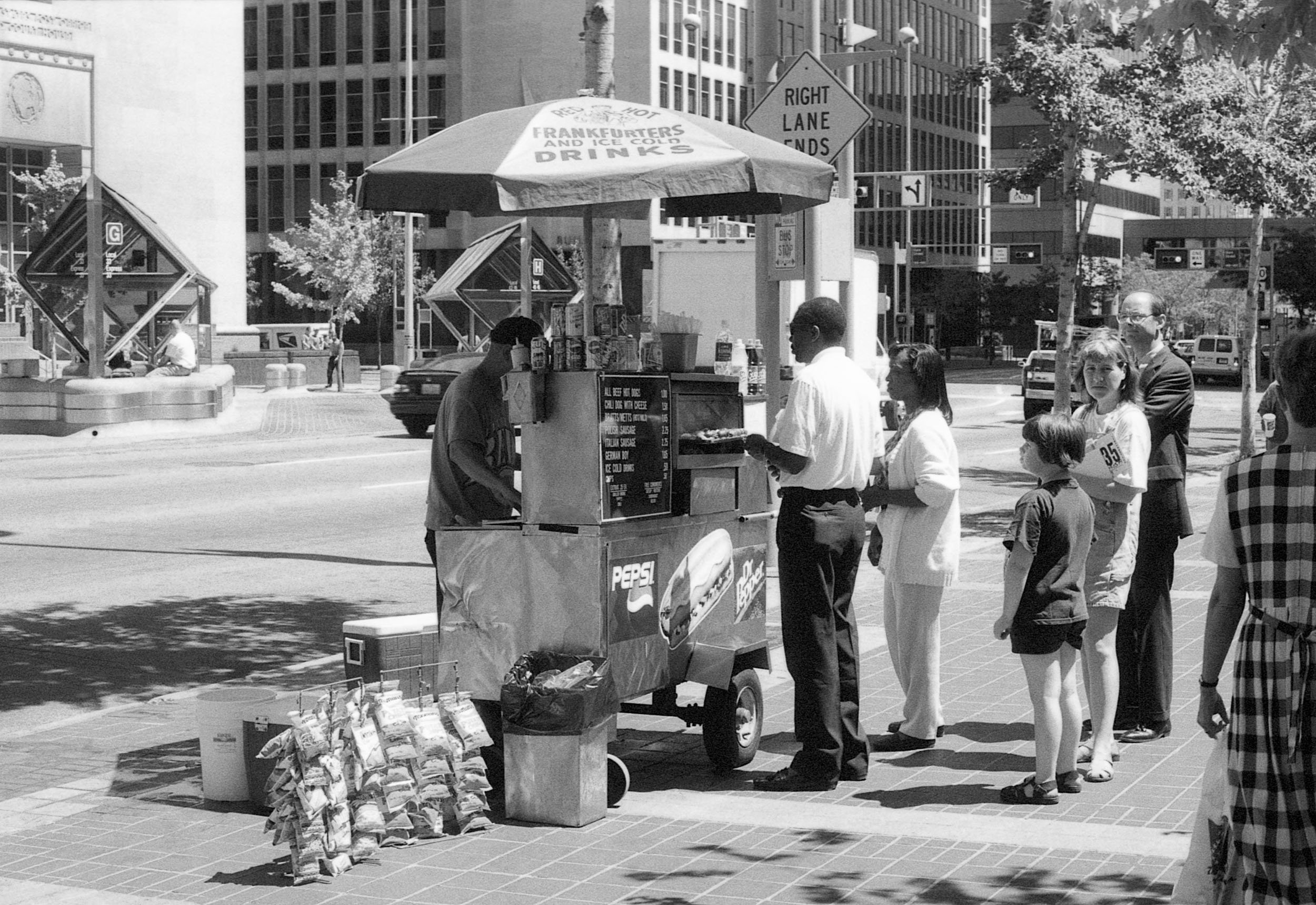 A black-and-white photo of a street scene with a food cart selling drinks and snacks. People are standing in line in front of the cart, including a man in a white shirt, a woman, and children. There are buildings, trees, and cars in the background.