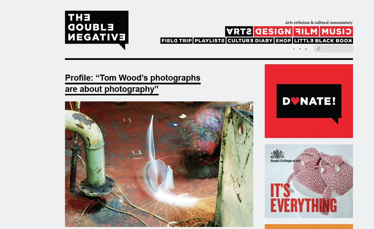 A webpage with a header titled "The Double Negative" and a main article about Tom Wood's photographs, featuring an image of a welding or metalworking process with sparks and bright light, surrounded by various navigation and advertisement elements on the webpage.