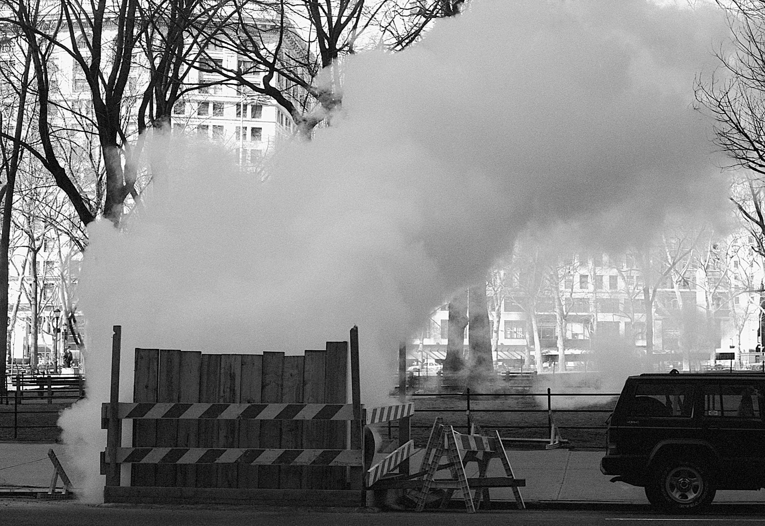 Construction site with large clouds of steam or smoke, wooden barriers, and a black SUV parked nearby, with trees and buildings in the background.