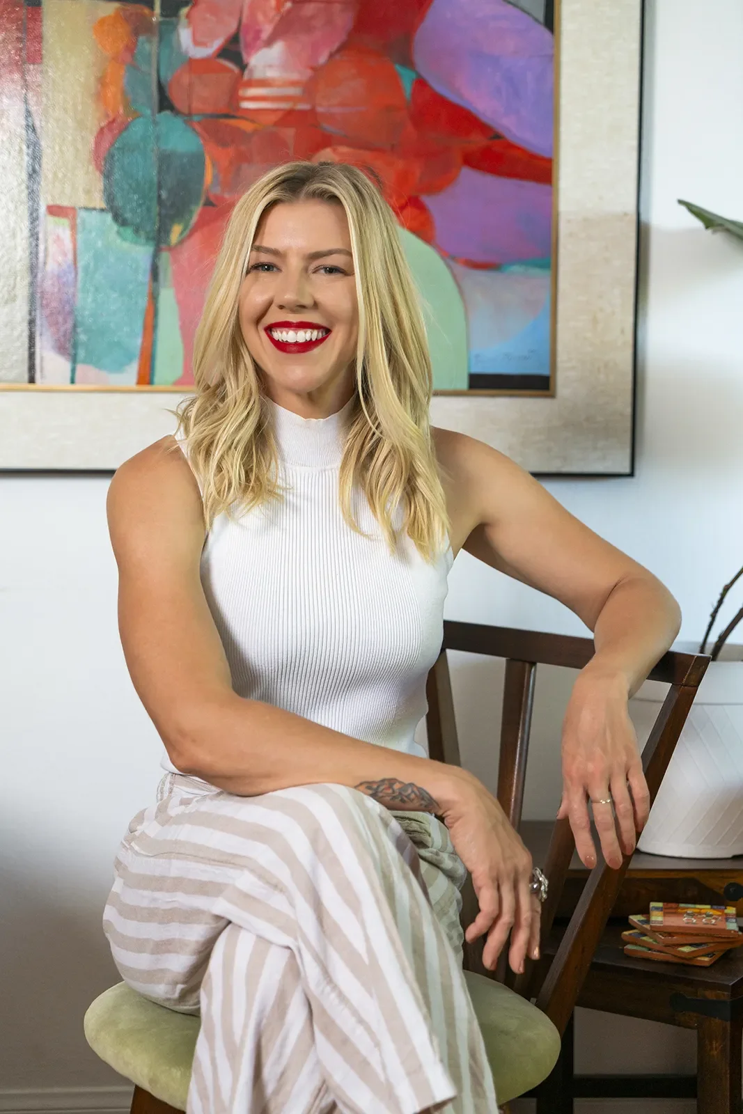 A woman with blonde hair, wearing a white sleeveless top and beige striped pants, sitting on a green cushioned chair, smiling with red lipstick, in a room with abstract colorful artwork and a potted plant in the background.