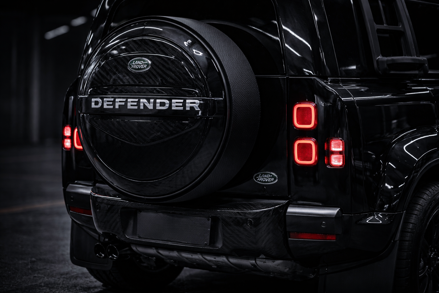 Rear view of a black Land Rover Defender with a spare tire cover and bright red tail lights in a dark indoor setting.
