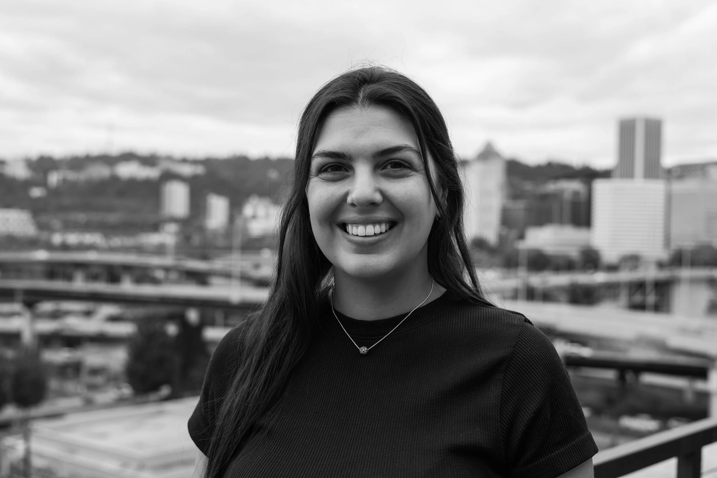 Close-up black-and-white portrait of a smiling young woman with long dark hair, standing outdoors with a cityscape in the background.
