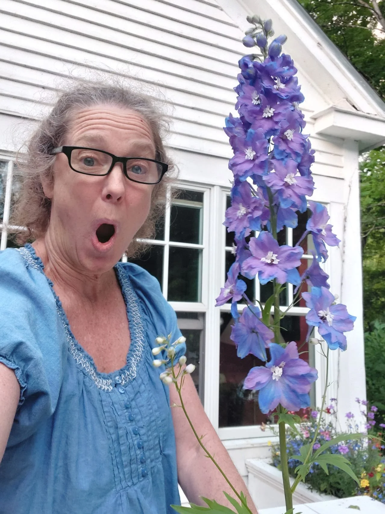 Surprised woman with glasses and purple flowers in front of white siding.