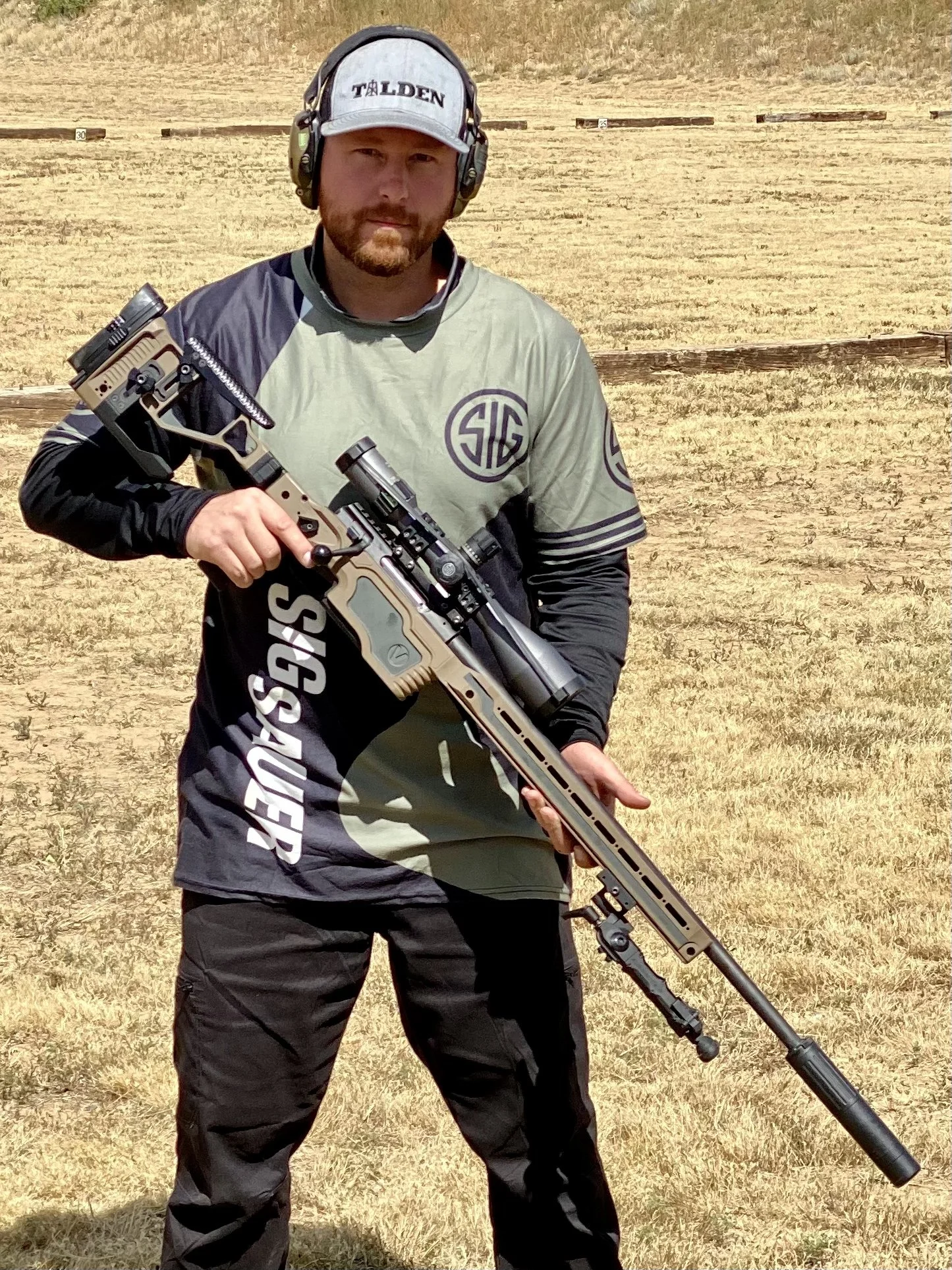 A man wearing a gray and black Taran tactical shirt, black pants, a white Talden hat, and hearing protection, holding a large sniper rifle with a scope in an outdoor shooting range with wooden targets in the background.