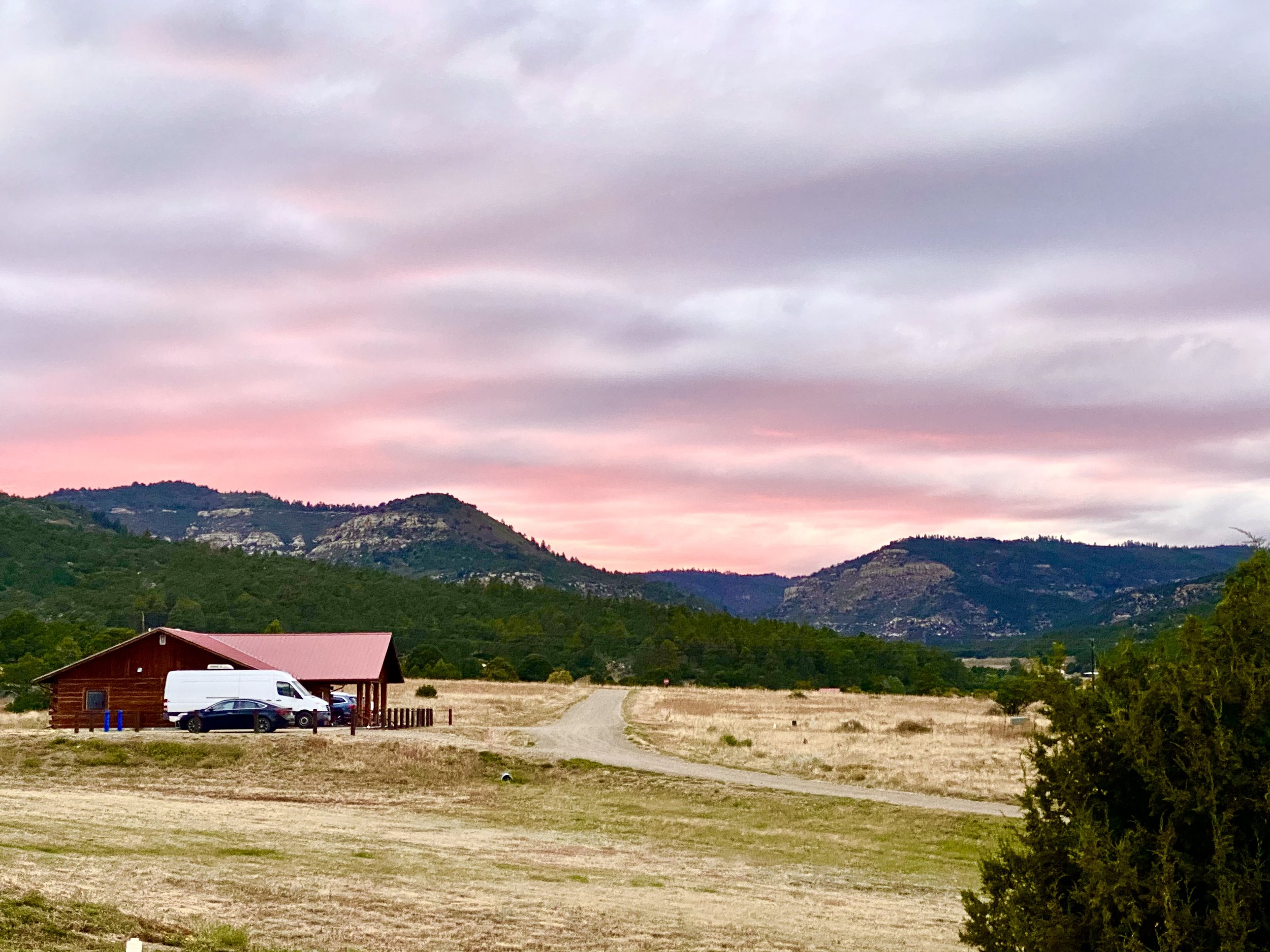 A rustic wooden building with a red roof surrounded by a few parked cars, set in a rural landscape with hills and a pink and purple sky at sunset.