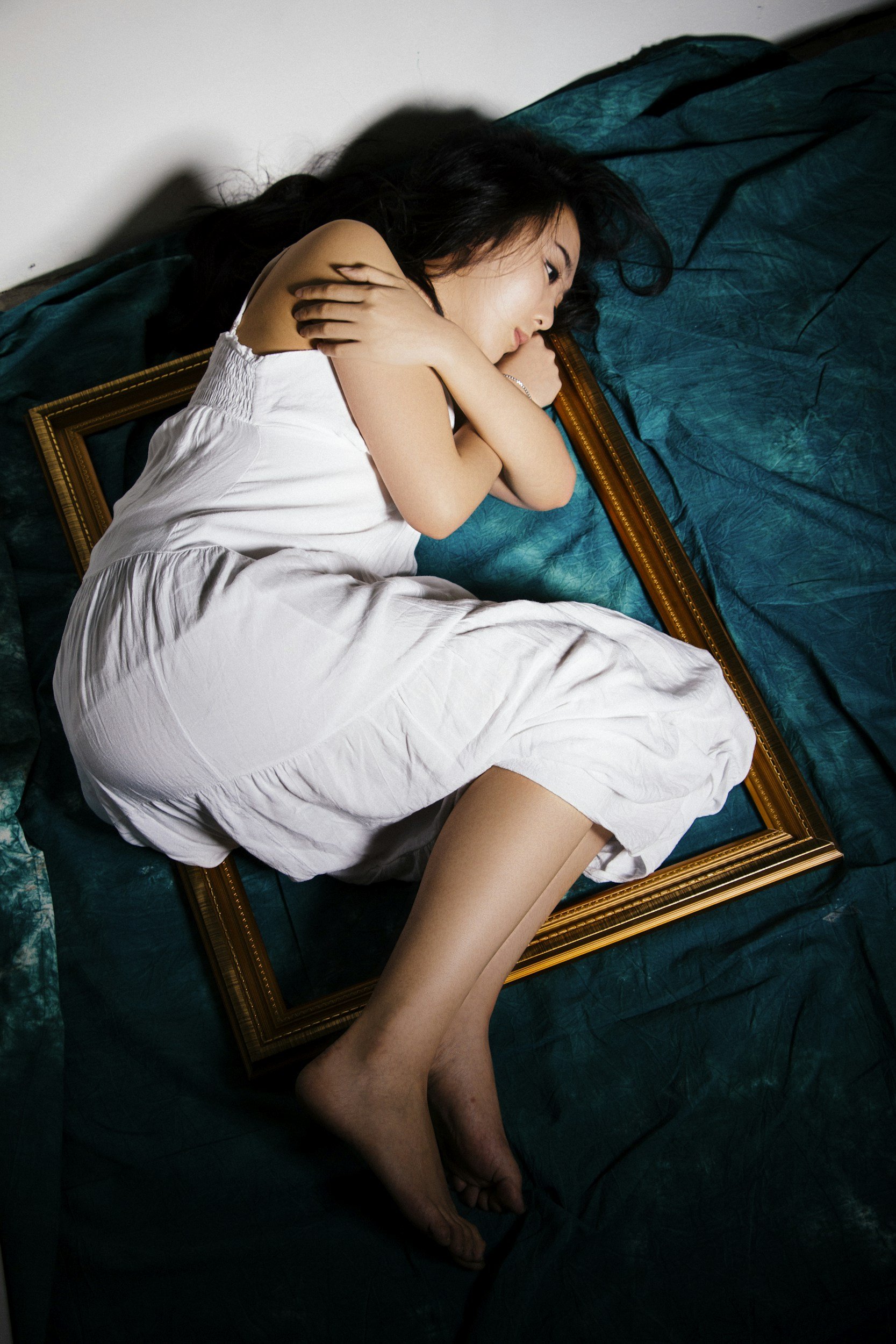 A woman sleeping on a bed with her head resting on a pillow, curled around a picture frame, wearing a white dress.
