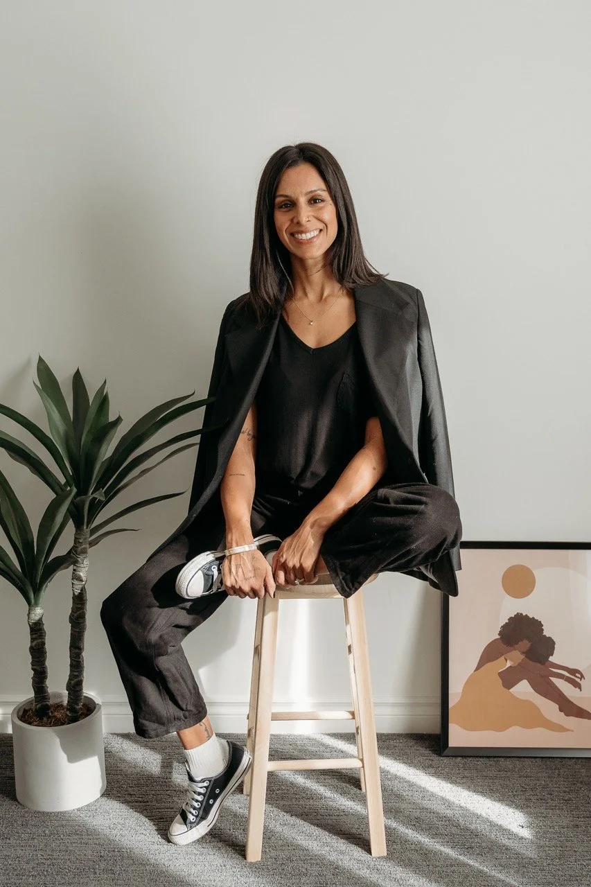 A woman sitting on a wooden stool in a room with a potted plant and a framed artwork. She is wearing a black blazer, black top, black pants, and black sneakers with white soles. She has shoulder-length dark hair and is smiling at the camera.