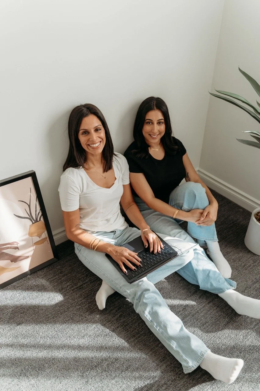 Two women sitting on the floor with a laptop, smiling at the camera, in a minimalistic room with a framed artwork and potted plant.