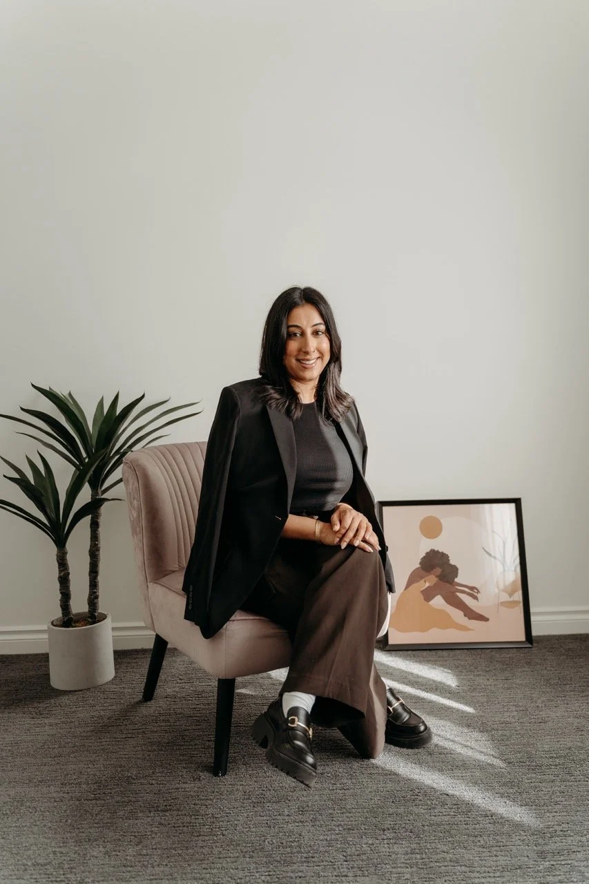 A woman sitting on a light pink chair in a room with gray carpet, smiling at the camera, wearing a black blazer, black shirt, brown pants, and black shoes. There is a potted plant to her left and a framed abstract artwork on the floor behind her.