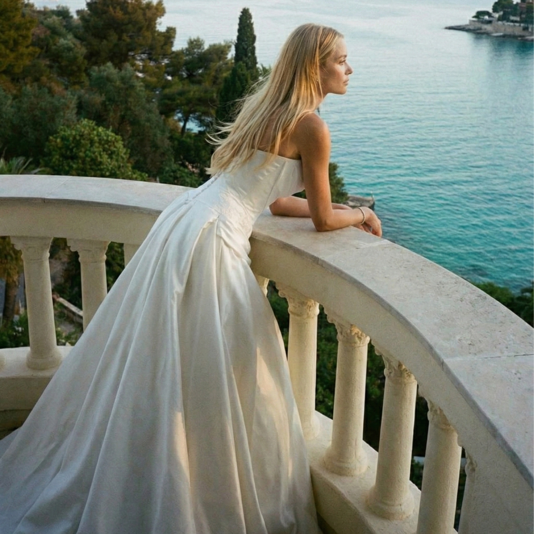 A woman in a white strapless dress leaning on a balustrade on an outdoor balcony, looking out over the water and trees.