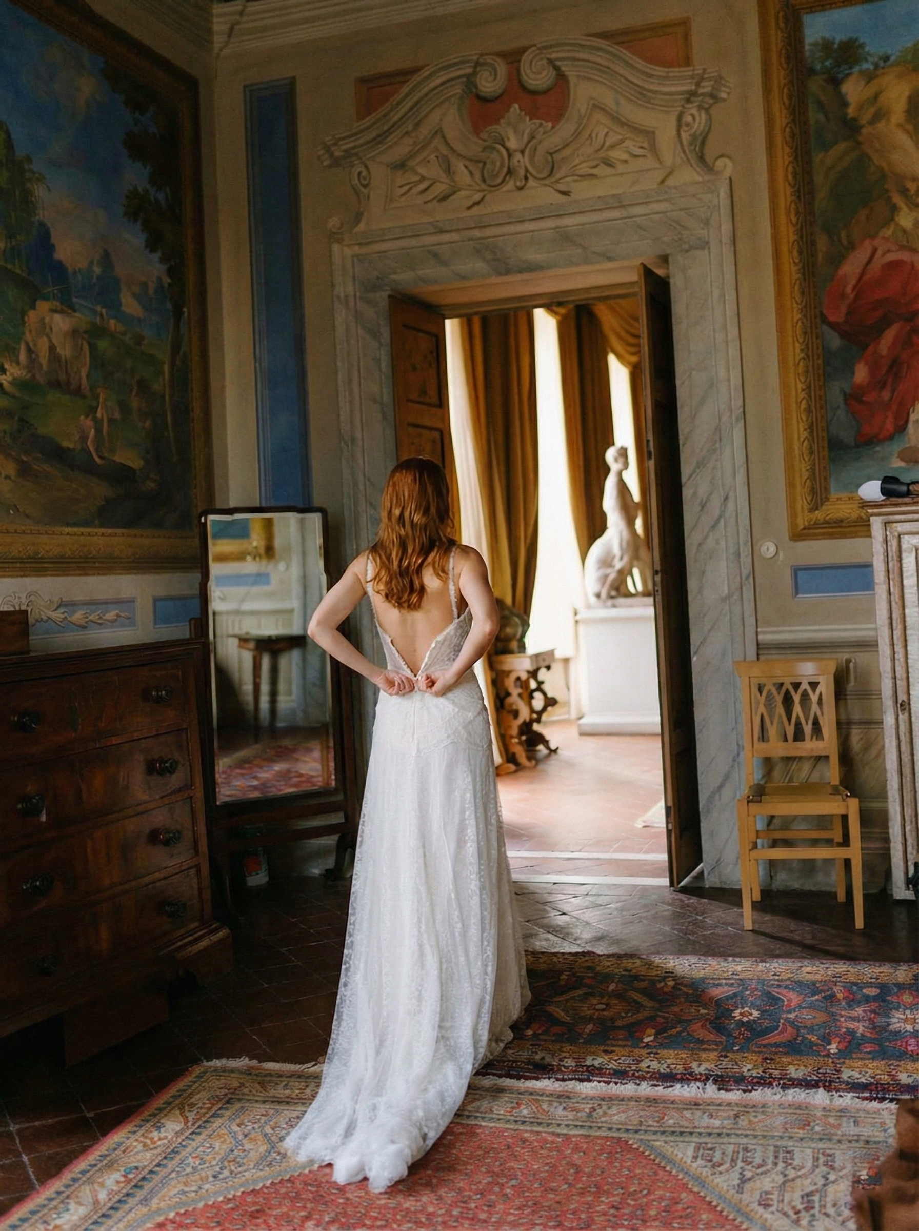 A woman with red hair wearing a white wedding dress, standing inside an elegant, ornately decorated room, looking through an open doorway towards a sunny room with curtains and a sculpture of a seated woman.