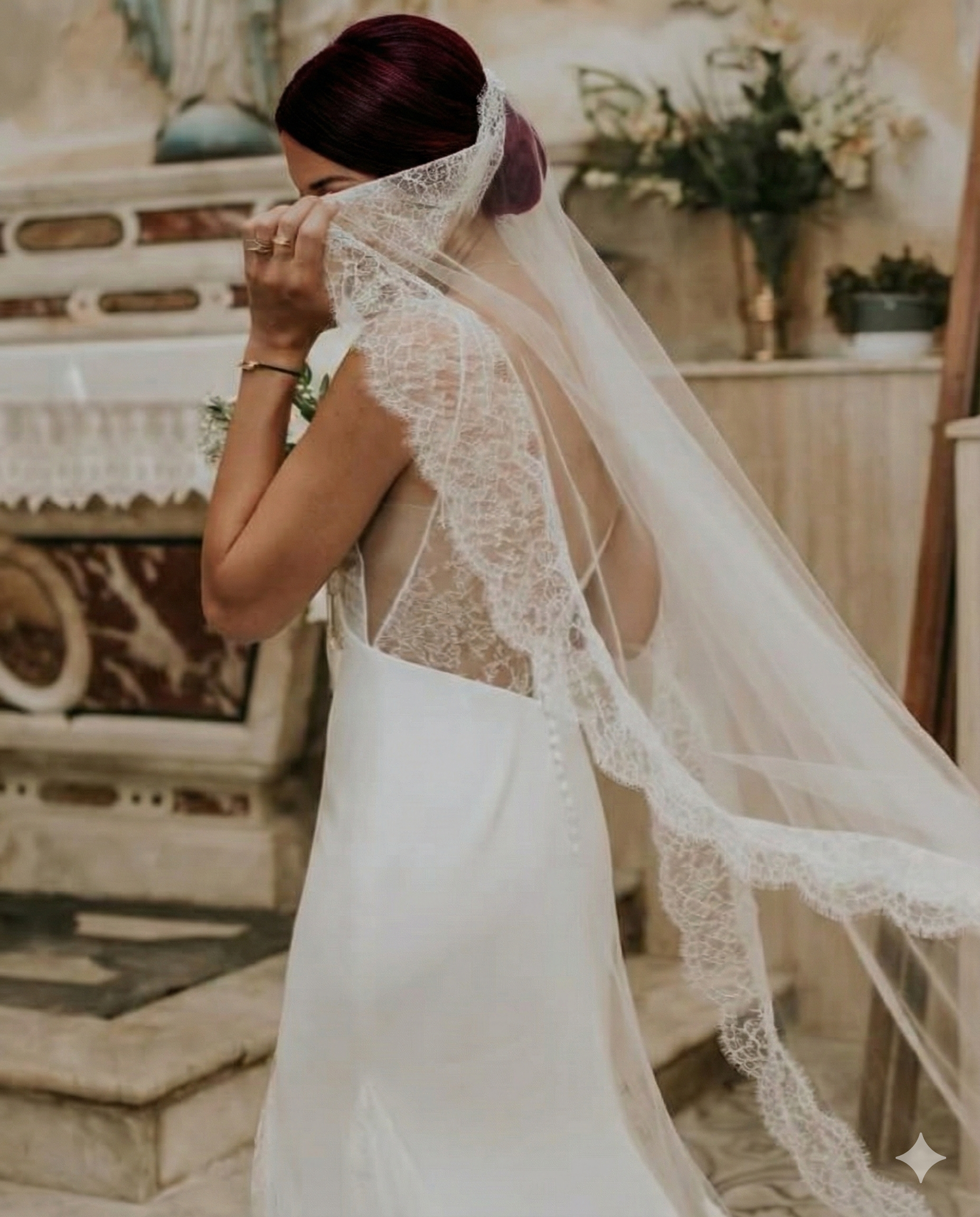 A bride in a wedding dress with lace details, holding a veil over her face, standing in a decorated room.