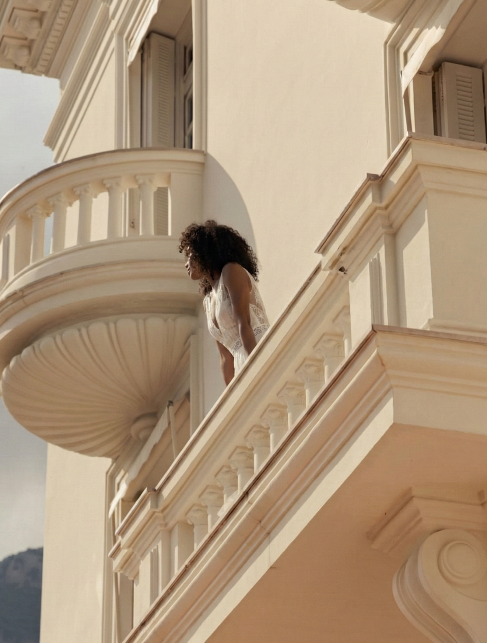 A woman with curly hair leaning on a balcony railing of a cream-colored building with ornate architectural details.