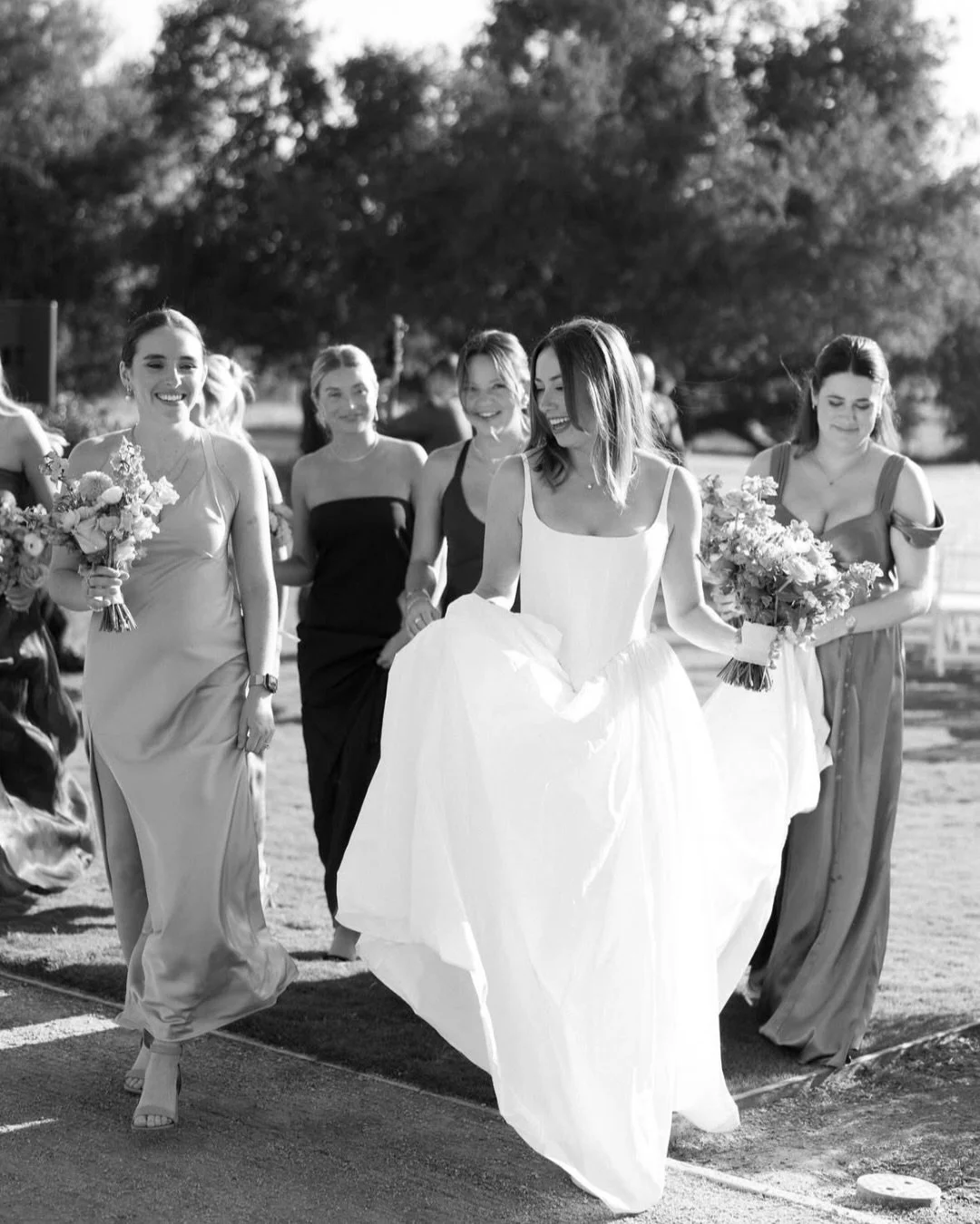 A group of women in elegant dresses walking outdoors, with the woman in the center wearing a white wedding gown and holding a bouquet, smiling and surrounded by bridesmaids.