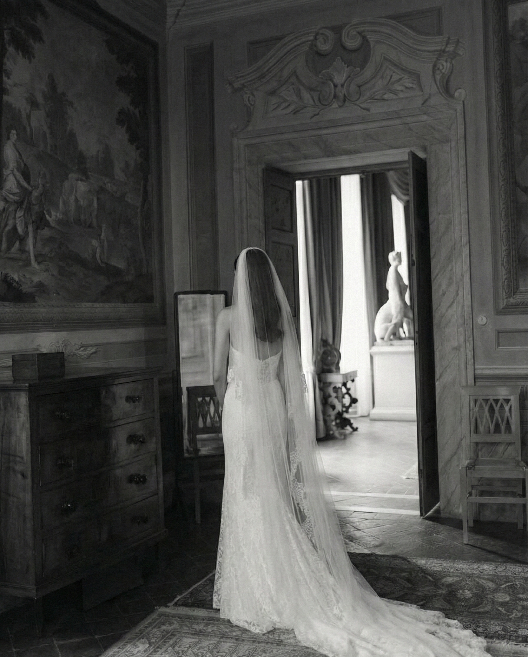 A bride in a wedding dress and veil standing inside an elegant room, facing away from the camera, looking towards a statue of a woman and a dog in a distinguished setting with drapes, paintings, and classic furniture.