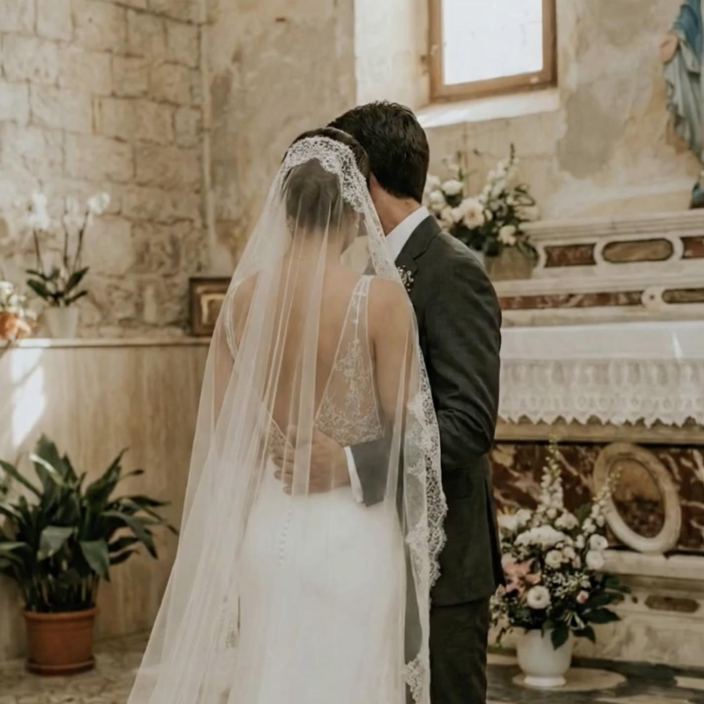 A bride and groom sharing a kiss during their wedding ceremony inside a chapel with stone walls and floral decorations.
