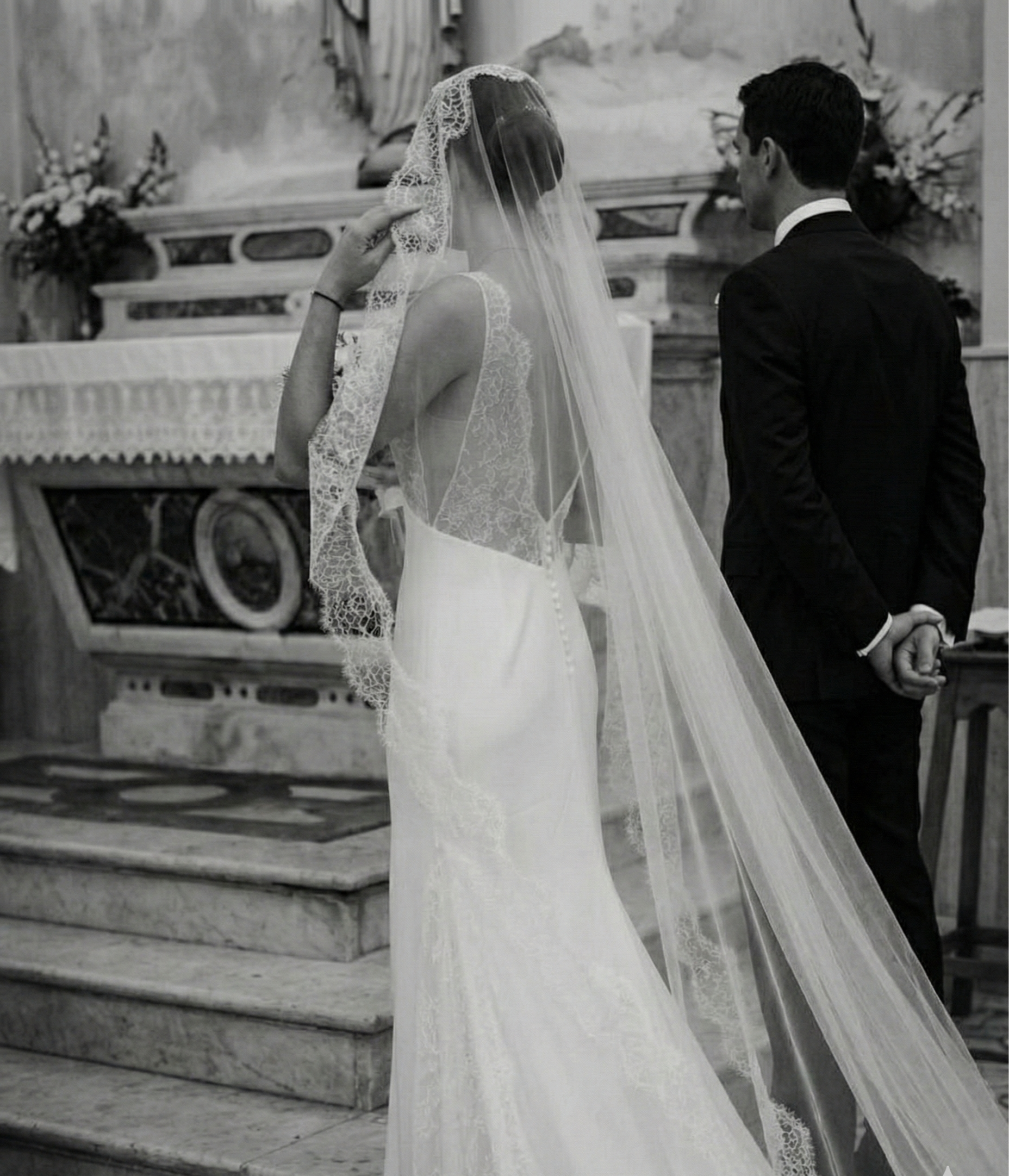 A bride and groom in a church during their wedding ceremony, with the bride wearing a lace wedding dress and veil, and the groom in a black tuxedo, standing with their hands clasped behind their backs.