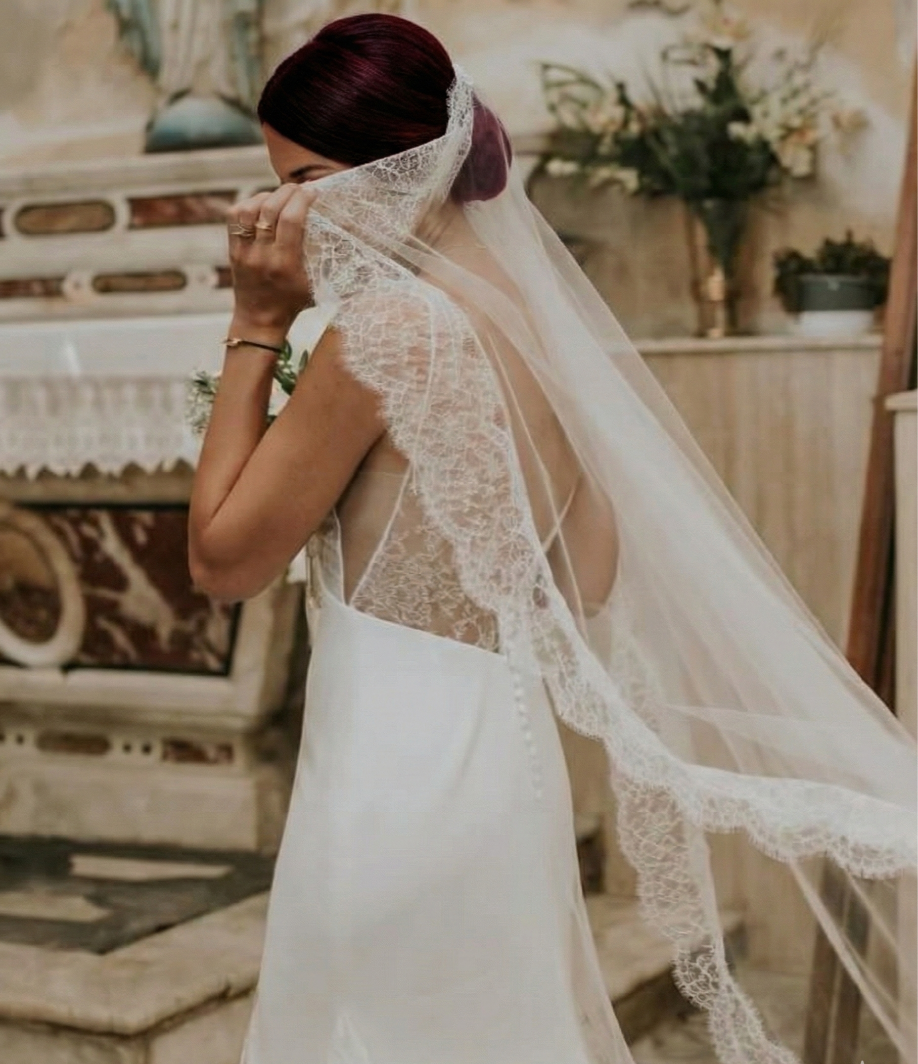 A bride in a wedding dress is holding and adjusting her lace veil in a vintage-style room with floral decorations.